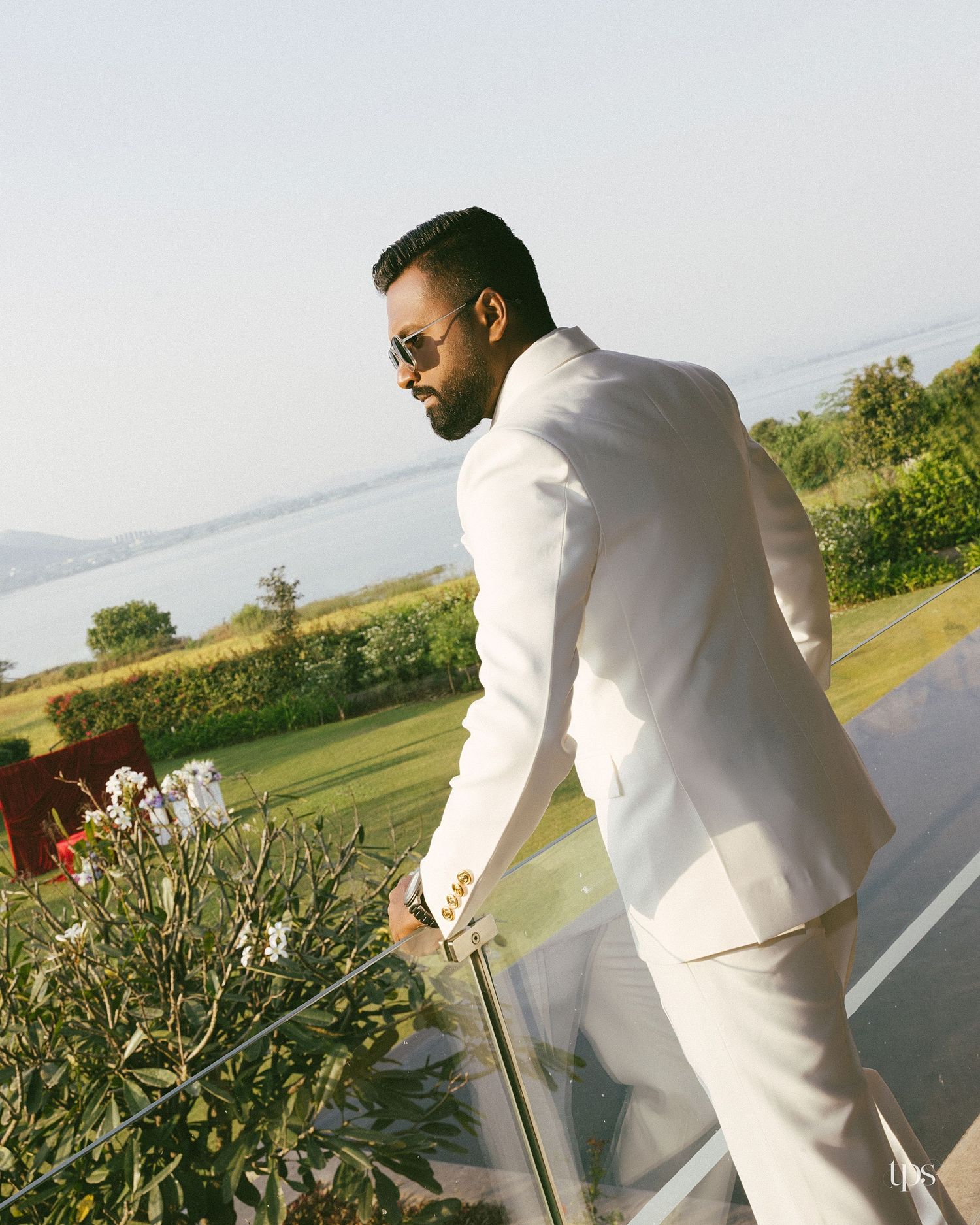 Man in a white suit and sunglasses stands at a glass railing, looking over a lush green lawn and a calm lake at sunset.