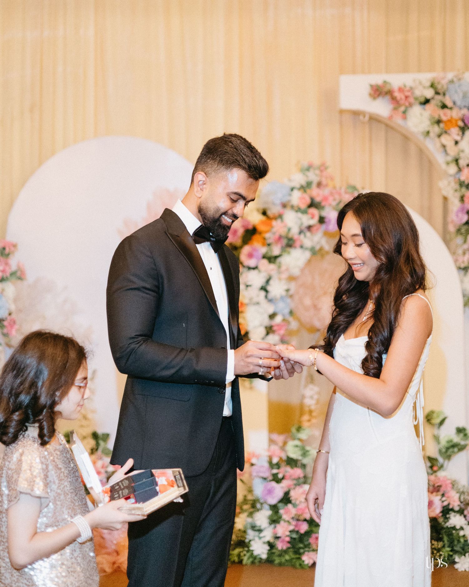Groom in black tuxedo exchanges rings with bride in white dress during a wedding ceremony as a young girl reads nearby, with floral decor.