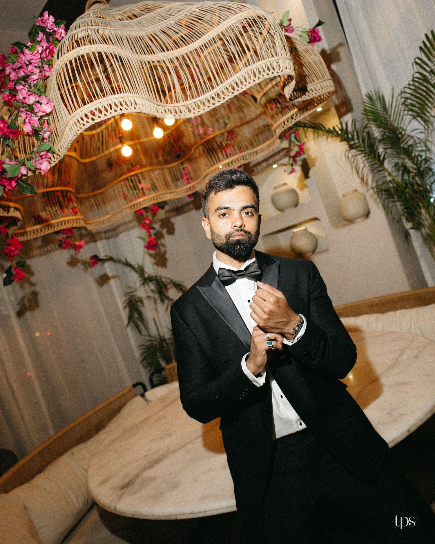 Groom in a black tuxedo and bow tie adjusts his cufflink beside a marble table under a woven rattan chandelier with pink floral accents.