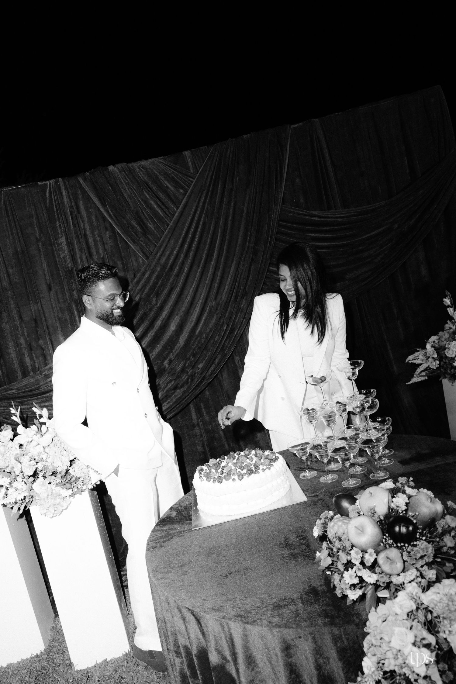 Black and white photo of a couple in white suits beside a frosted cake and champagne glasses, framed by flowers and dark velvet drapes.