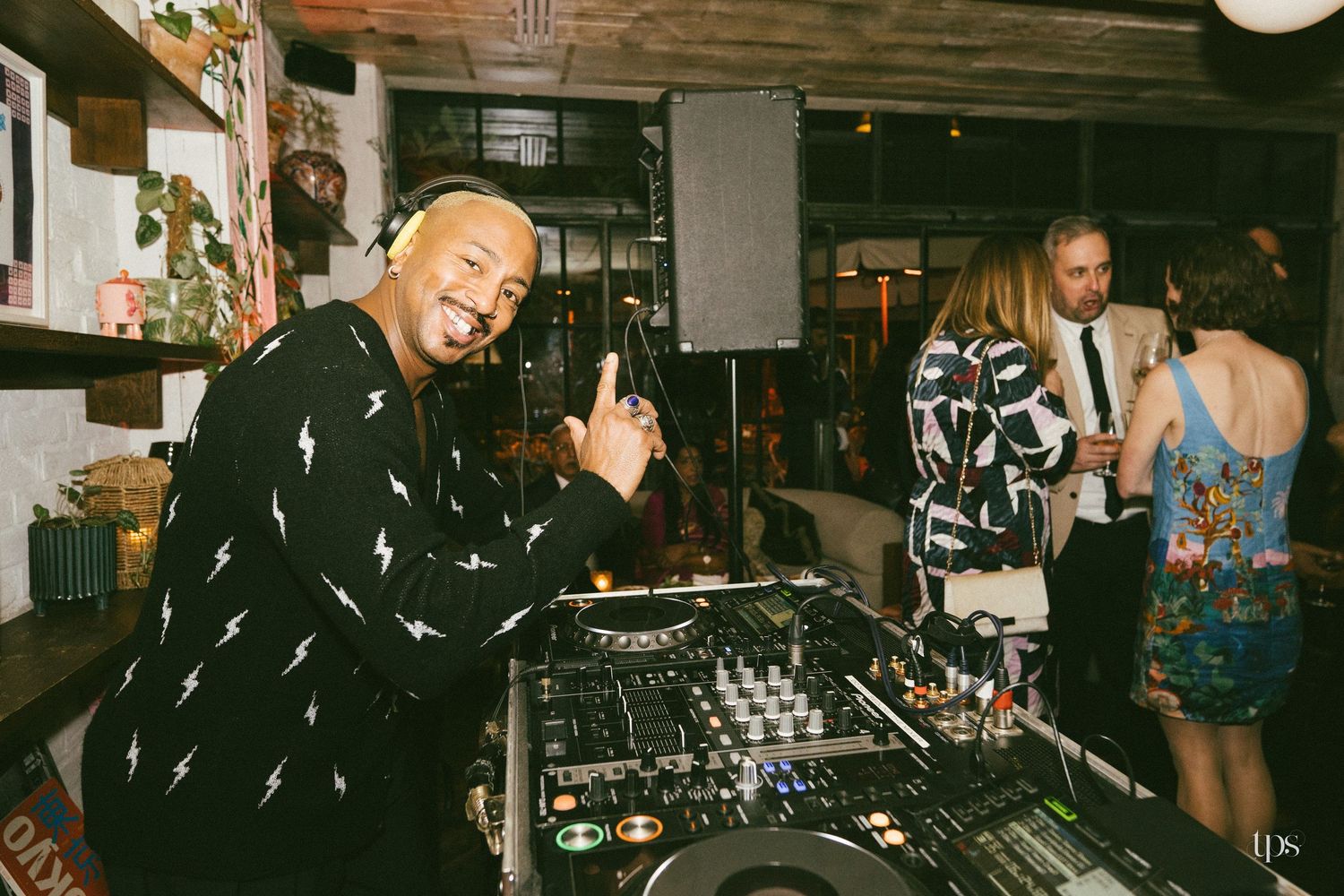 A DJ smiles at the camera behind a mixer at an indoor party with guests in patterned outfits in the background.