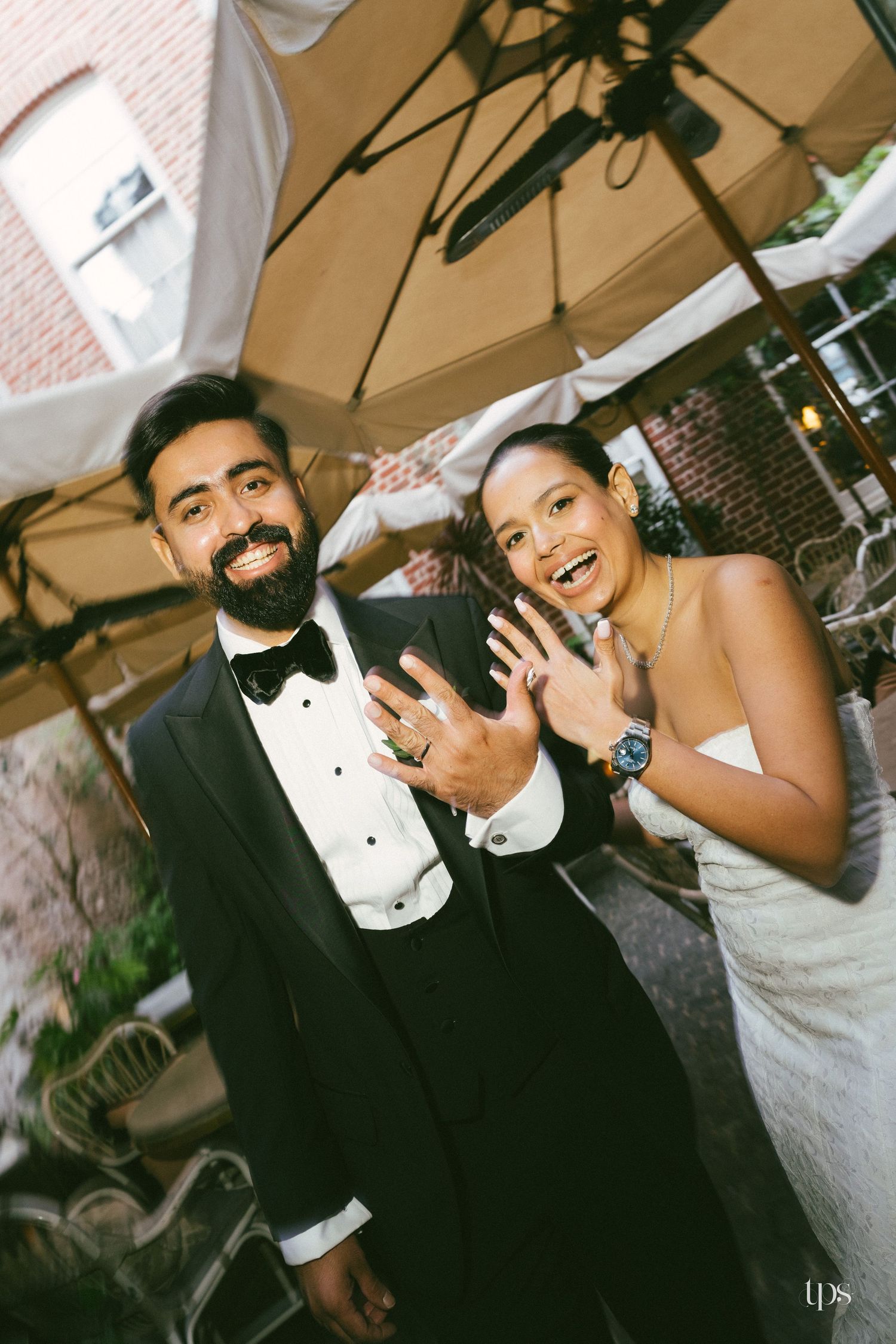 Smiling bride and groom in tuxedo showing wedding rings outdoors under umbrella at elegant reception.
