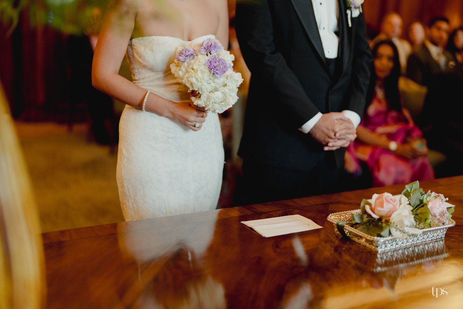 Bride in white gown holding purple and white bouquet stands with groom in tuxedo at wedding ceremony altar.