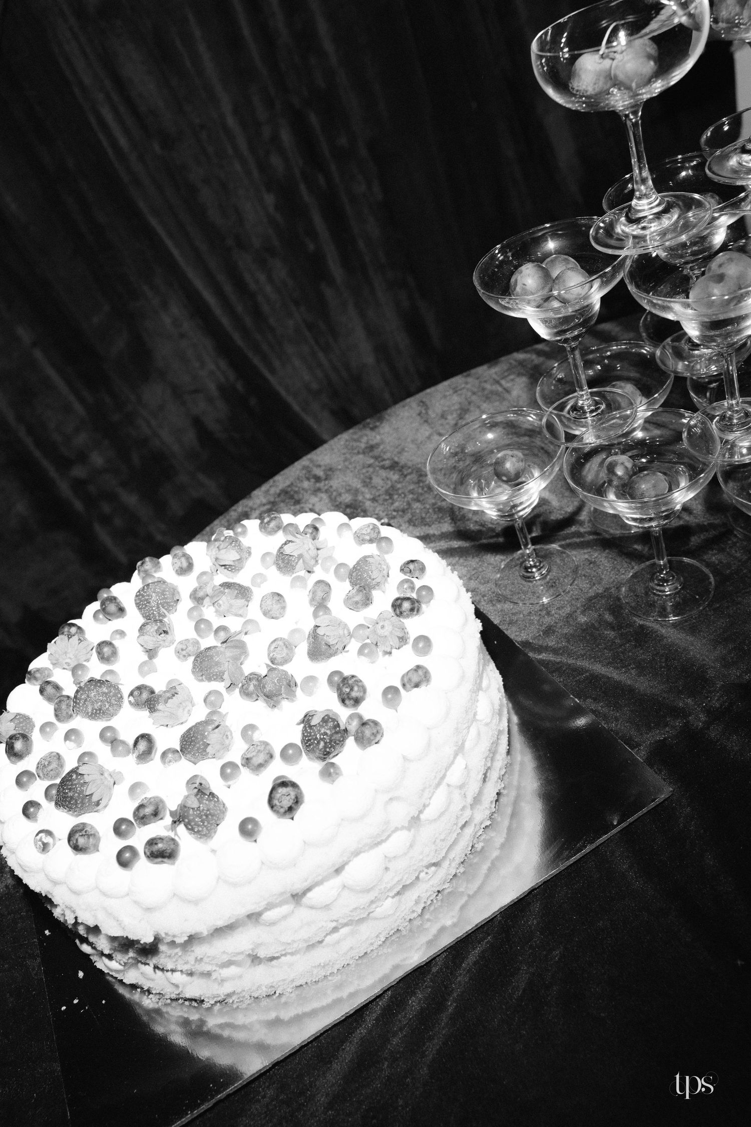 Black and white photo of a layered wedding cake topped with berries beside a champagne coupe glass tower on a draped table.