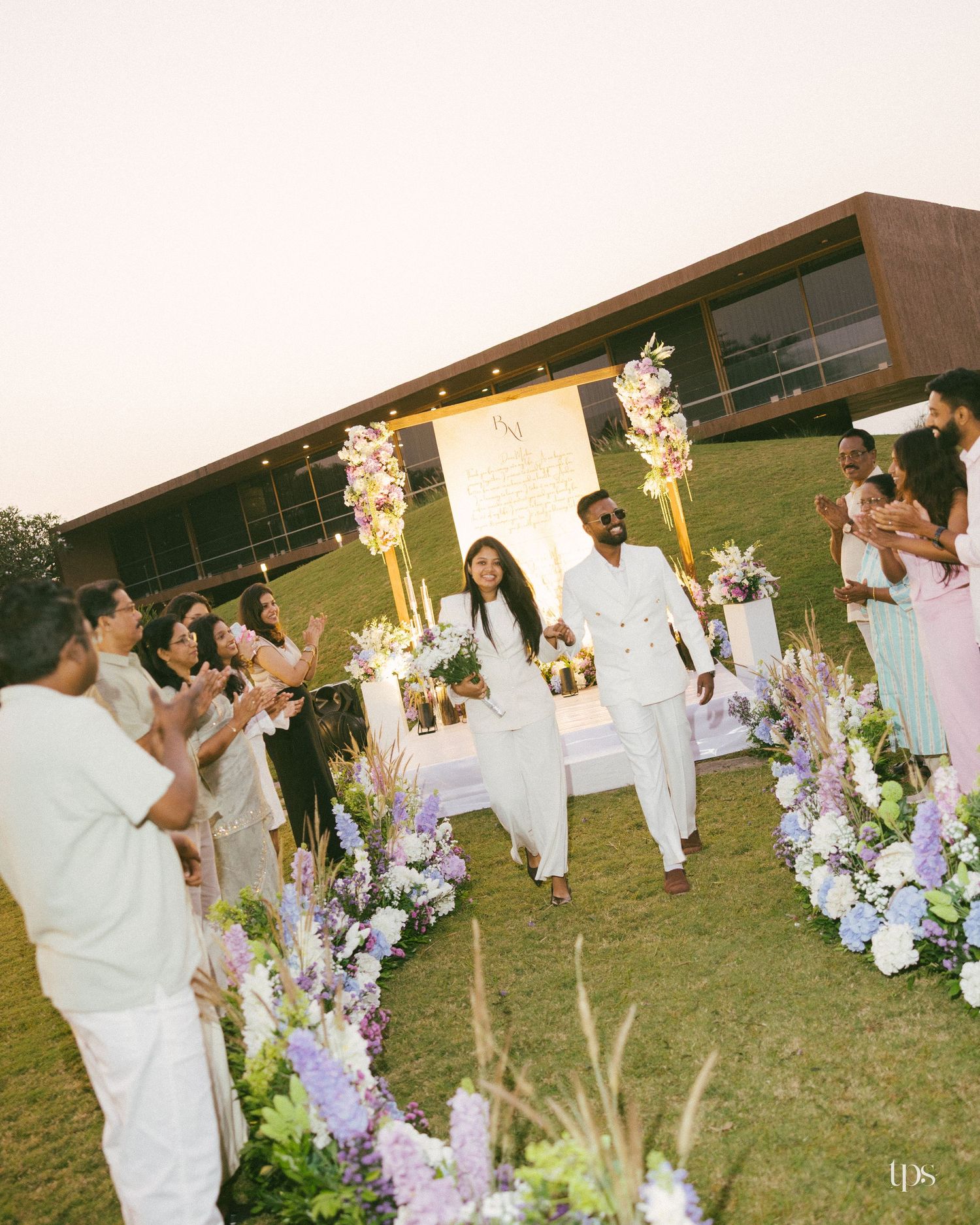 Couple in white outfits walk down a grass aisle lined with lavender and white flowers as guests applaud at an outdoor wedding.