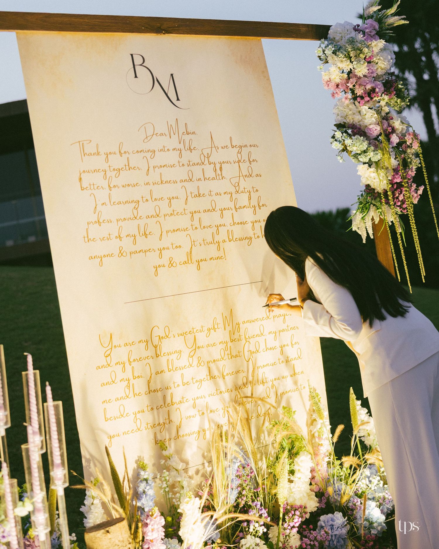 Woman in white writes on a large cream wedding message banner with BM monogram, framed by pastel flowers and candles at an outdoor ceremony.