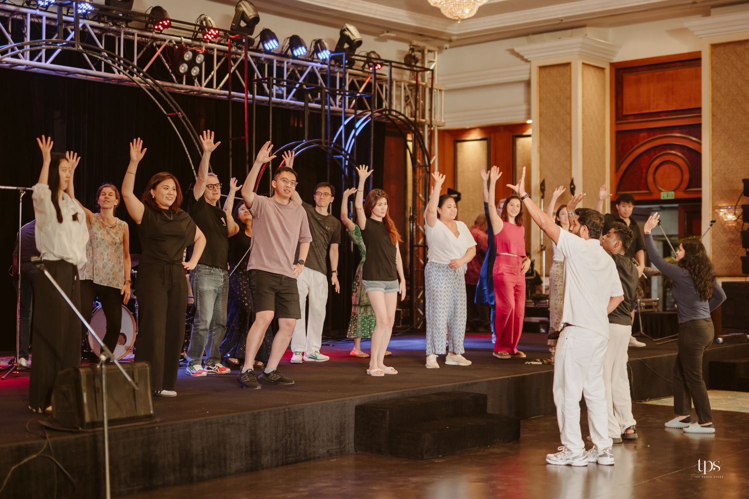 Group of performers on stage with hands raised during a theatrical rehearsal in an elegant ballroom venue.
