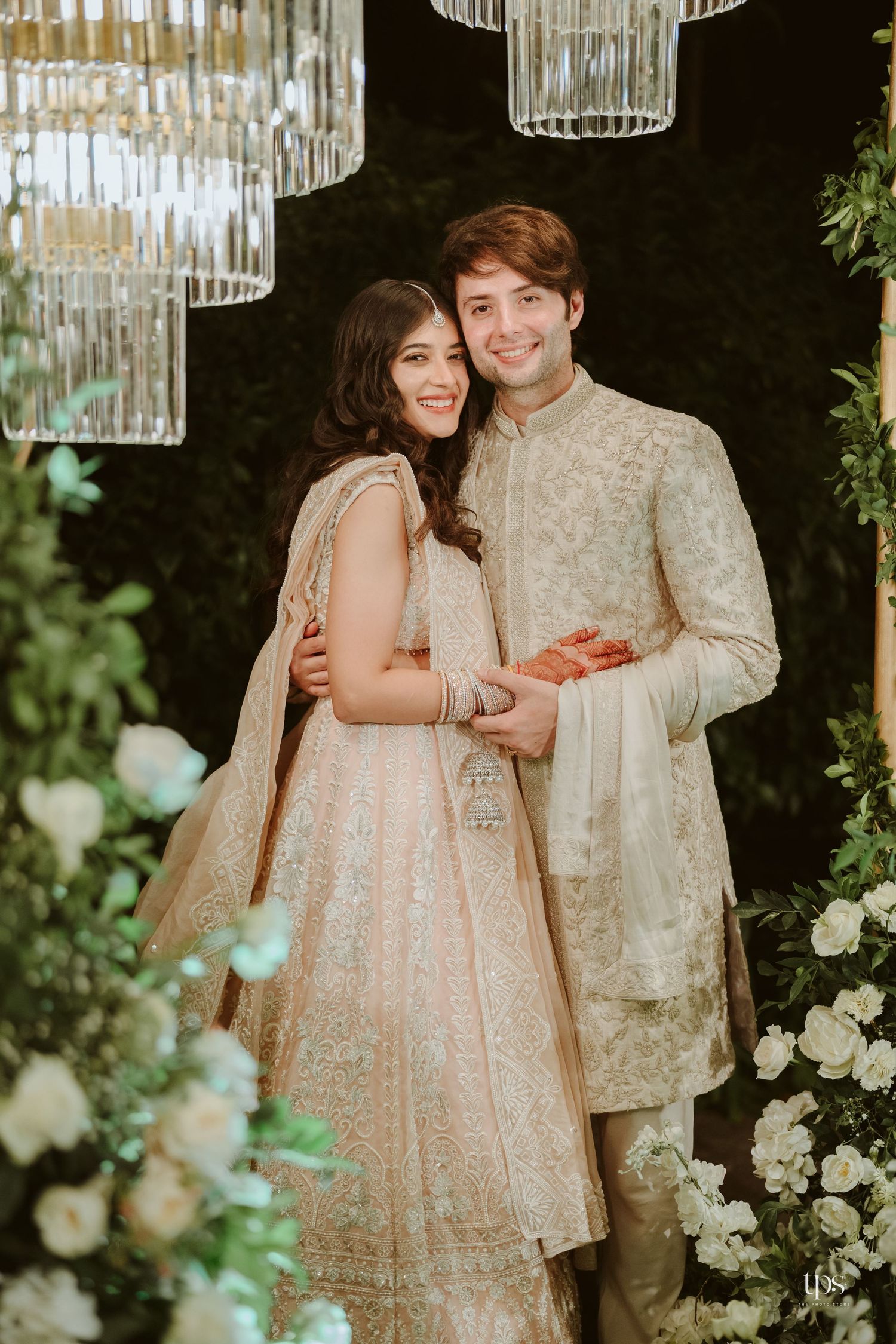 Couple in traditional Indian wedding attire, woman in embroidered lehenga and man in sherwani, posing at elegant floral decor.