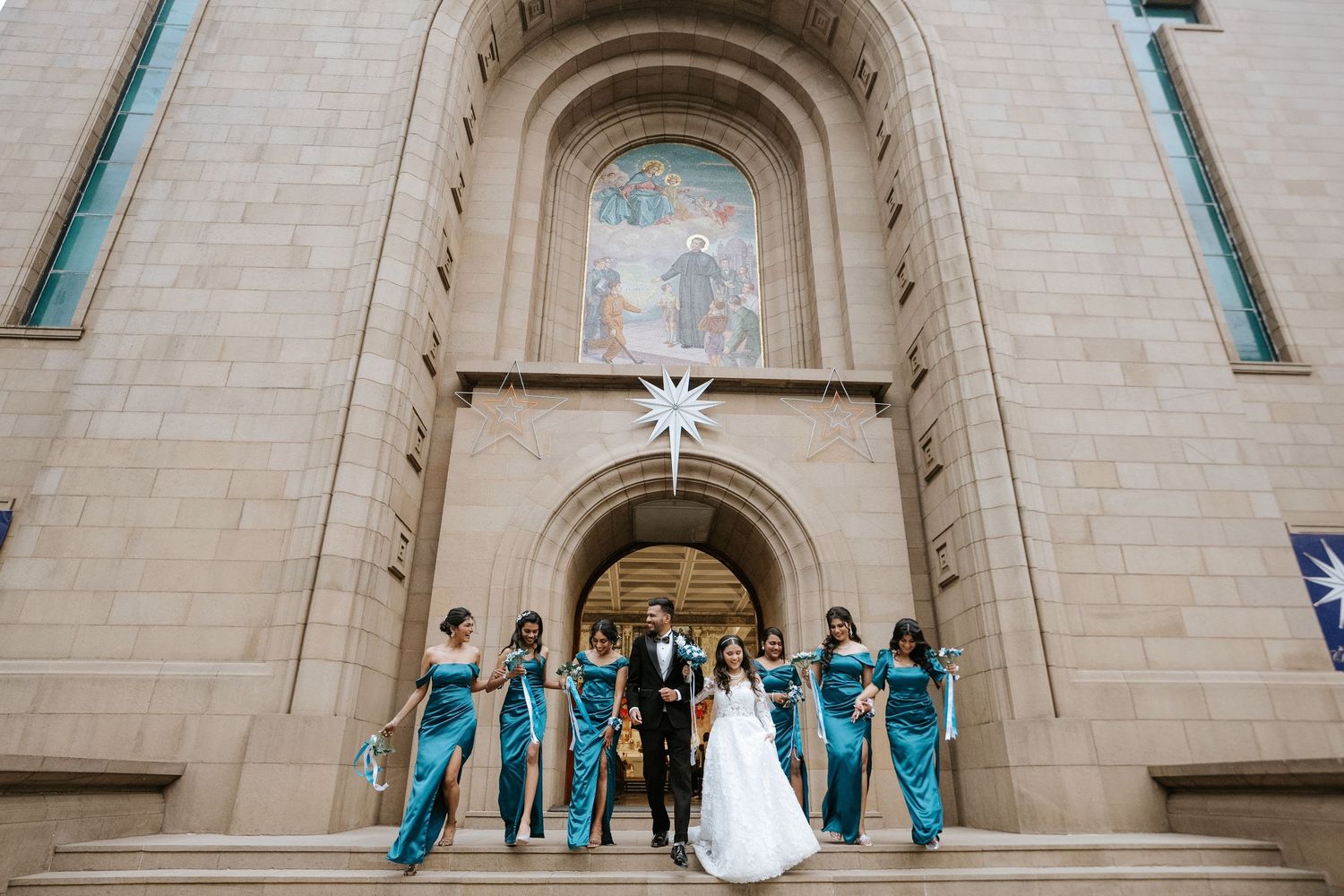 Wedding party posing before ornate stone church architecture with teal bridesmaid dresses, captured by a wedding Photographer in Mumbai.