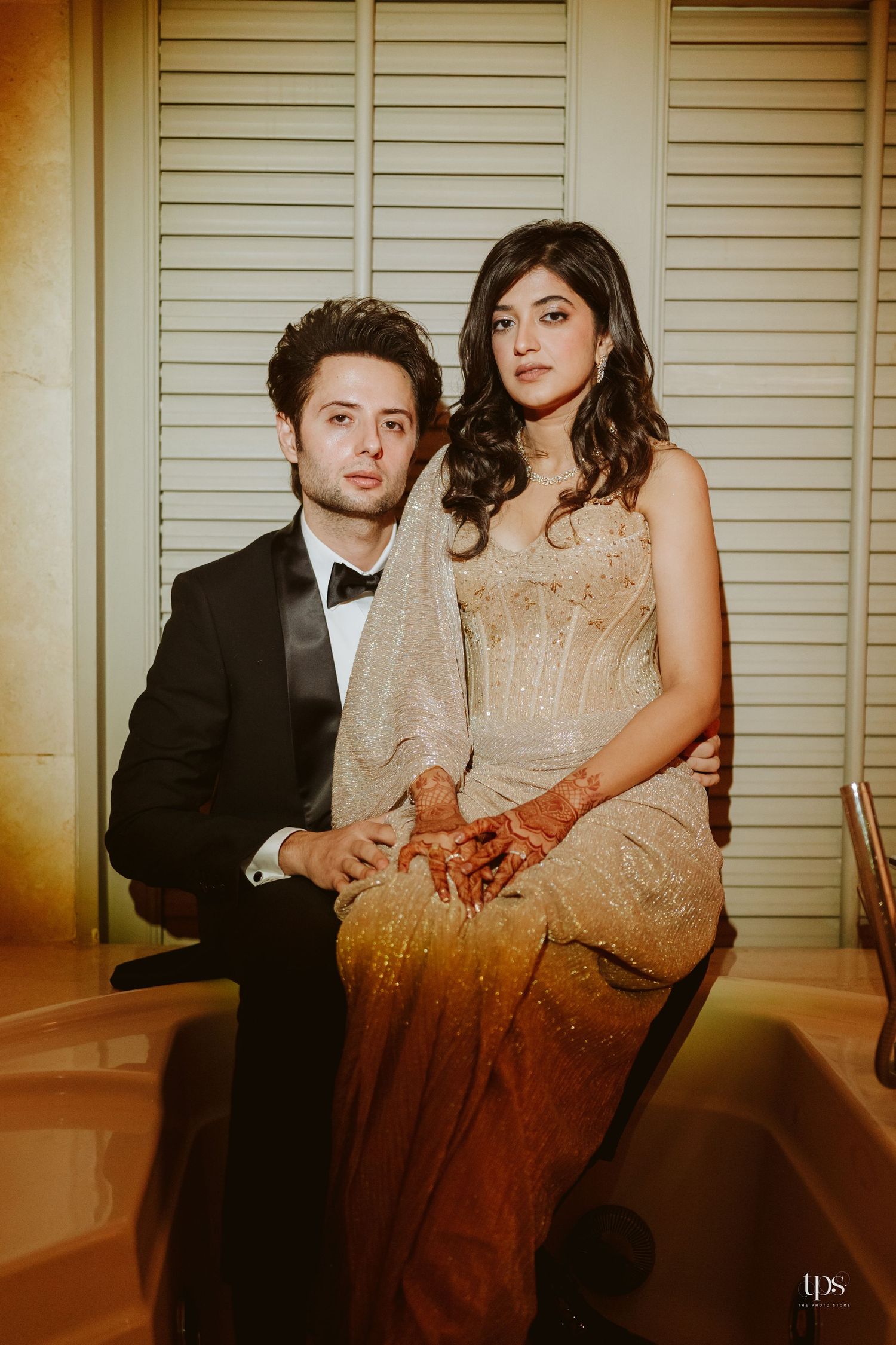 Couple in elegant attire posing in a bathtub, man in tuxedo and woman in gold gown with mehndi.