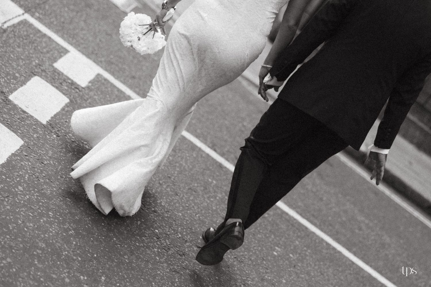 Black and white photo of a bride in white gown and groom in suit walking together on a crosswalk.