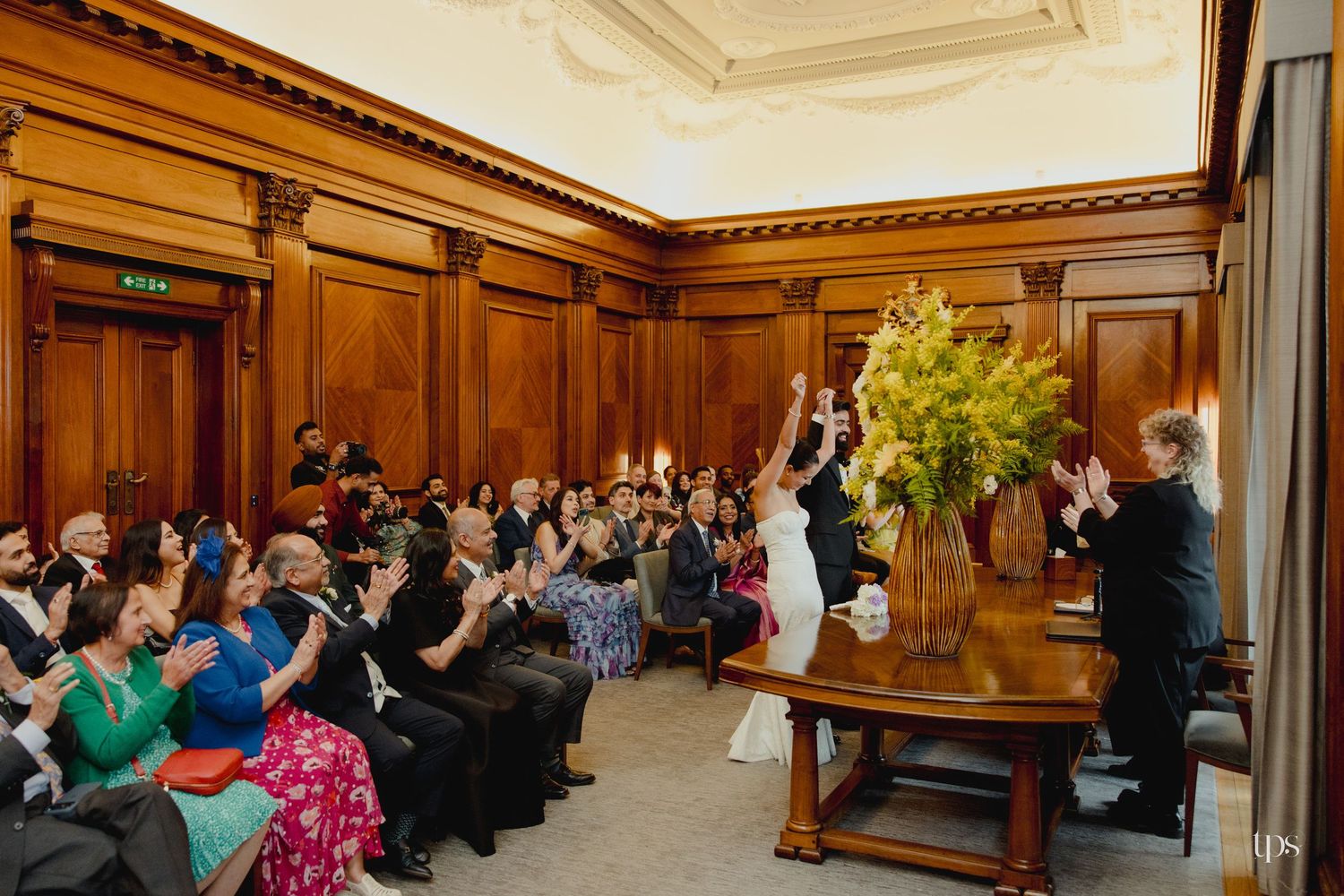 Joyful wedding ceremony in an elegant wood-paneled room with guests applauding as couple celebrates at the altar.
