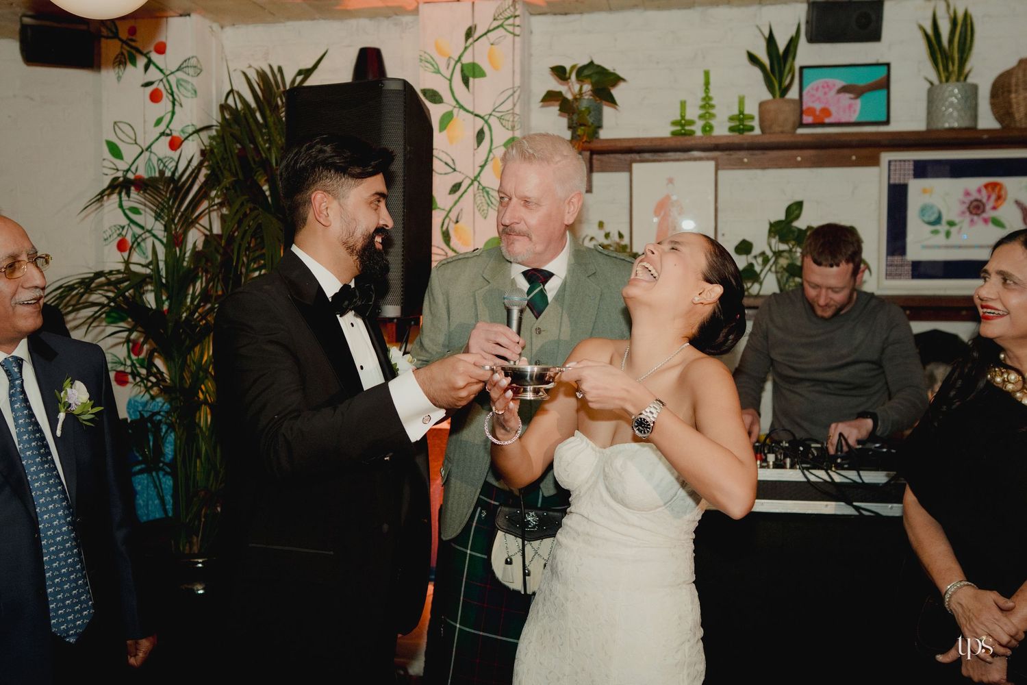 Bride and groom toasting with guests at wedding reception, man in tartan kilt, surrounded by lush green plants.