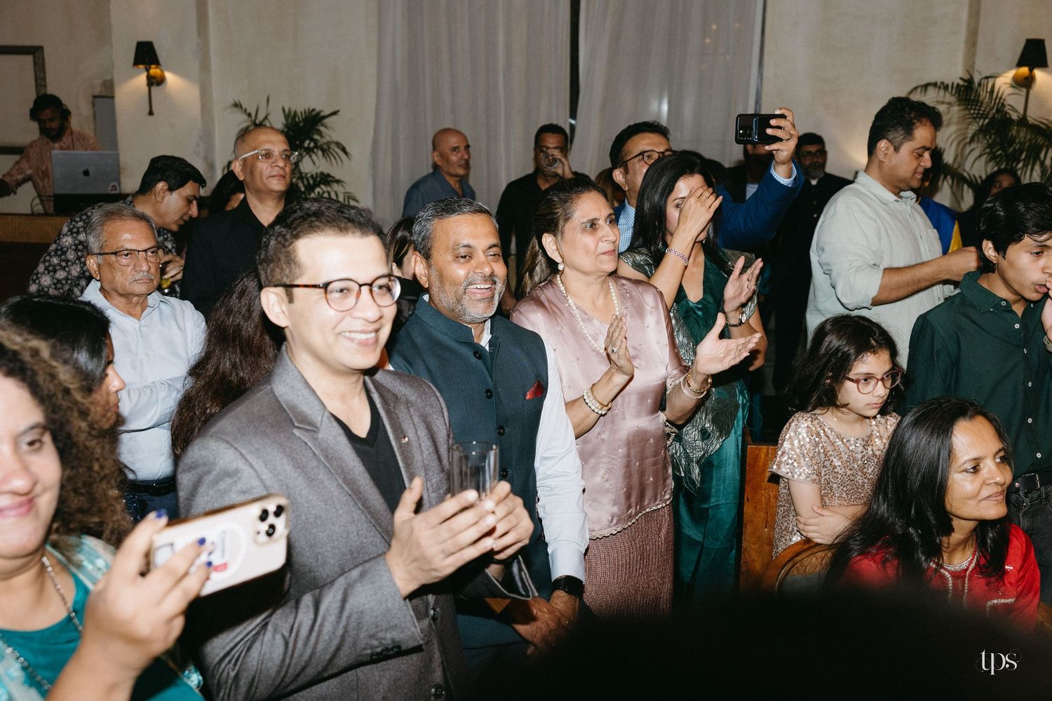 A smiling crowd of guests applauds and films with phones at an indoor event, with a man in a gray blazer and glasses clapping in front.