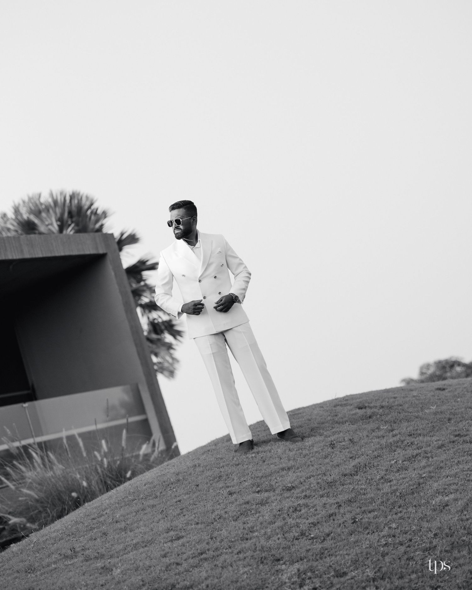 Man in sunglasses and a white double-breasted suit stands on a grassy hill beside a modern concrete building in a black and white photo.