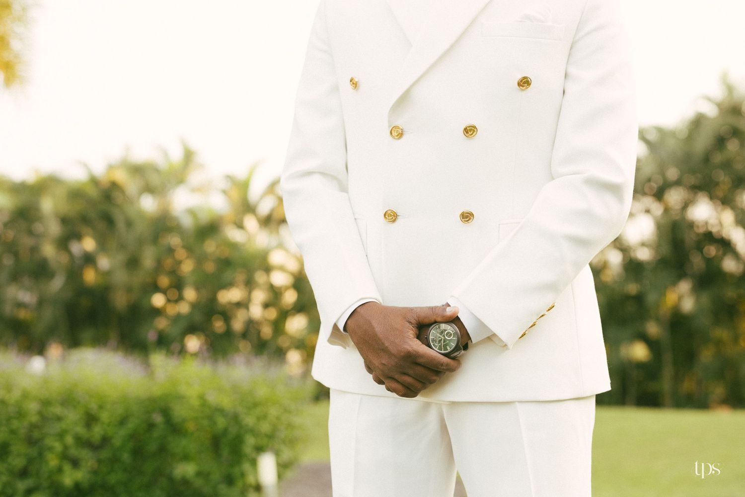 Groom in a white double-breasted suit with gold buttons clasps his hands in front of a lush tropical garden.