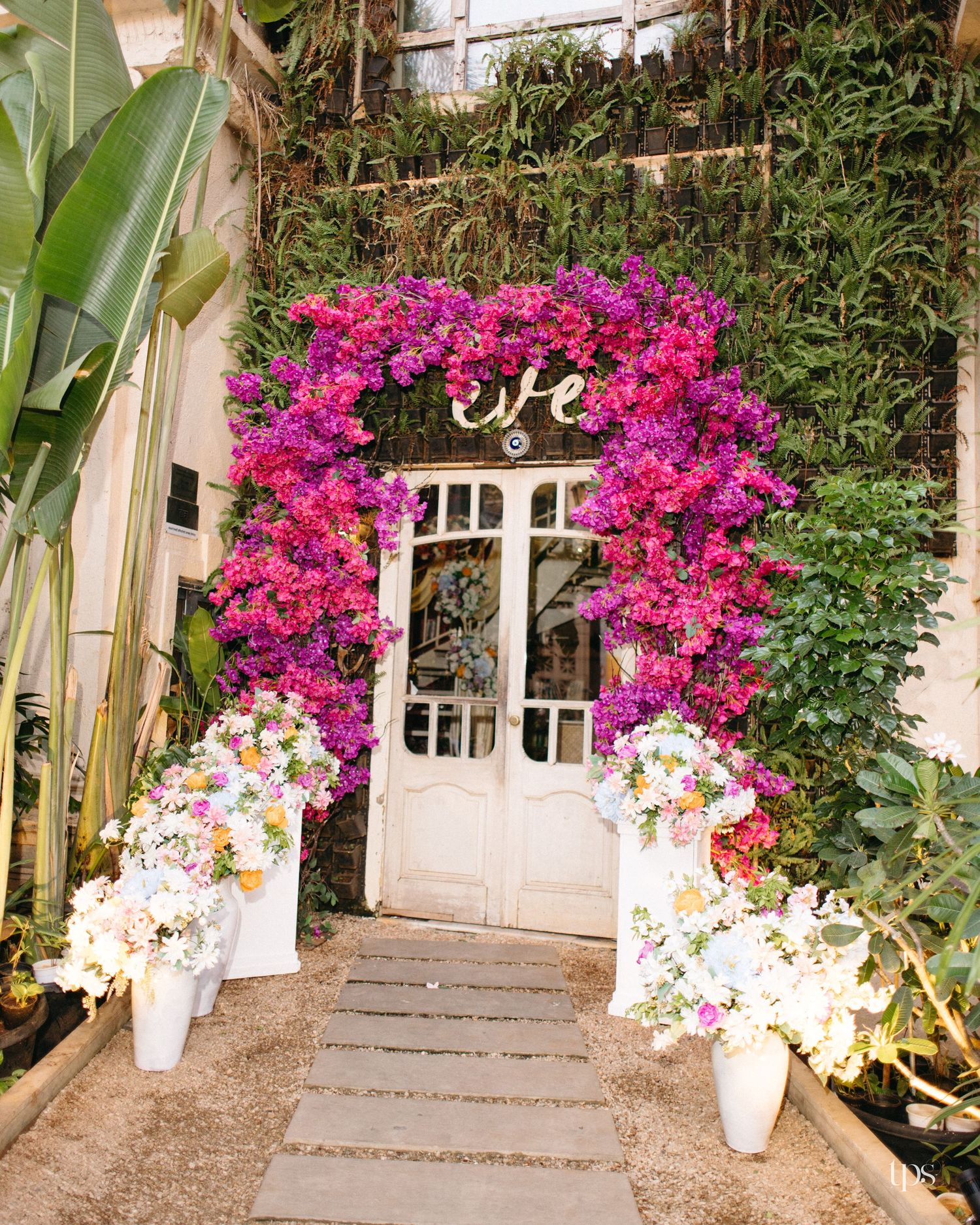 Vibrant purple bougainvillea arch over white French doors with a stone path and flower arrangements in white vases in a lush garden.