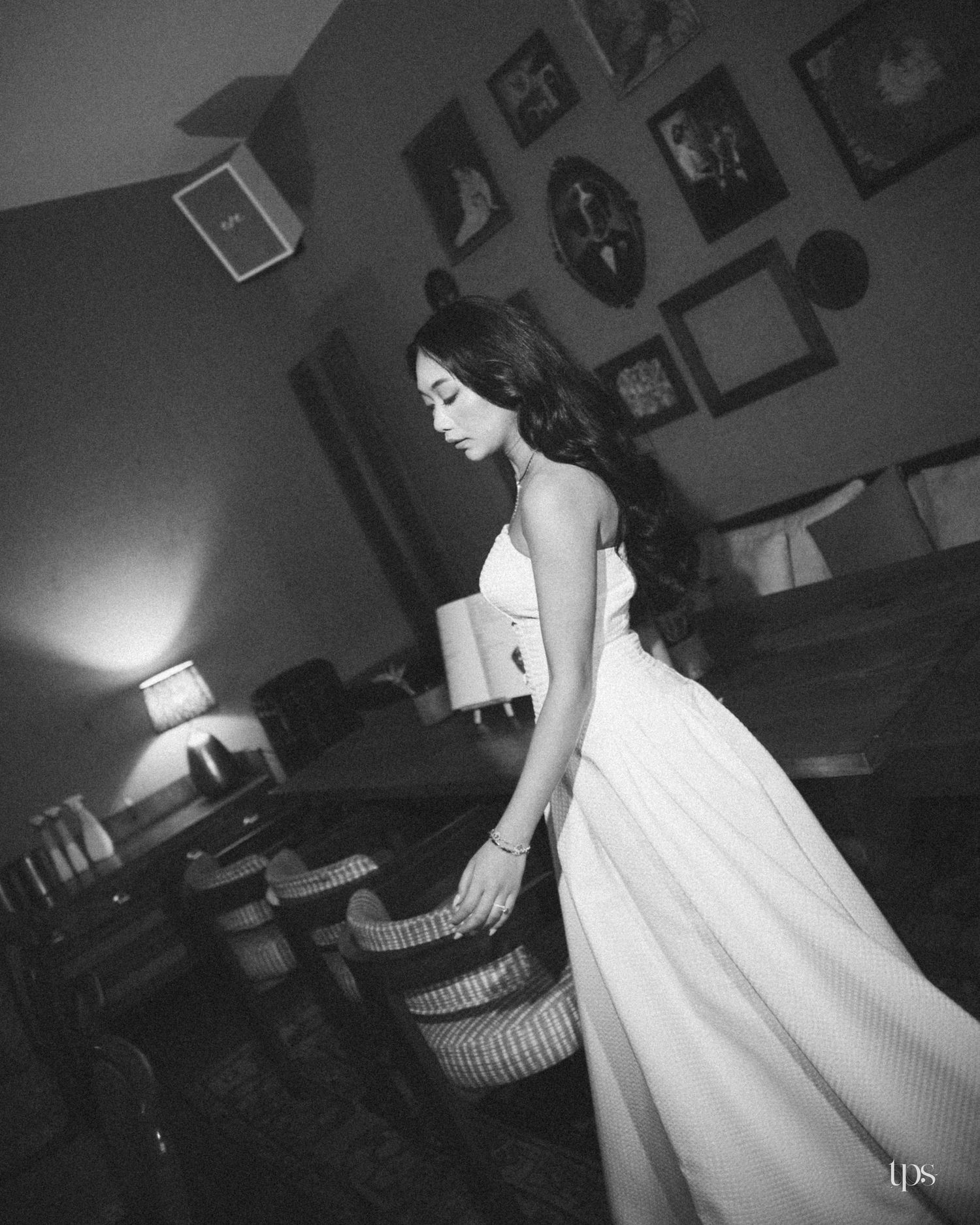 Black and white photo of a woman in a flowing white gown standing beside wicker chairs in a vintage room with framed wall portraits.