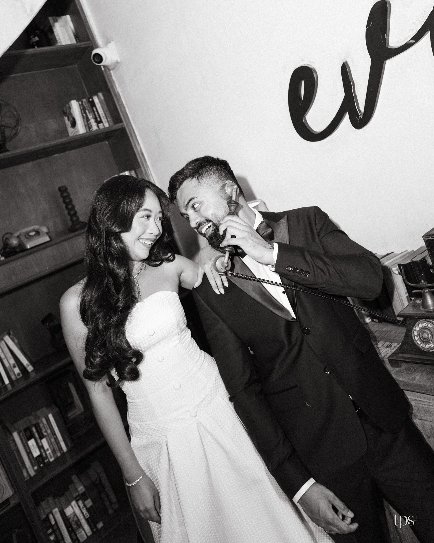 Black and white photo of a smiling bride in a strapless gown and a groom in a tuxedo laughing while holding a vintage telephone receiver.