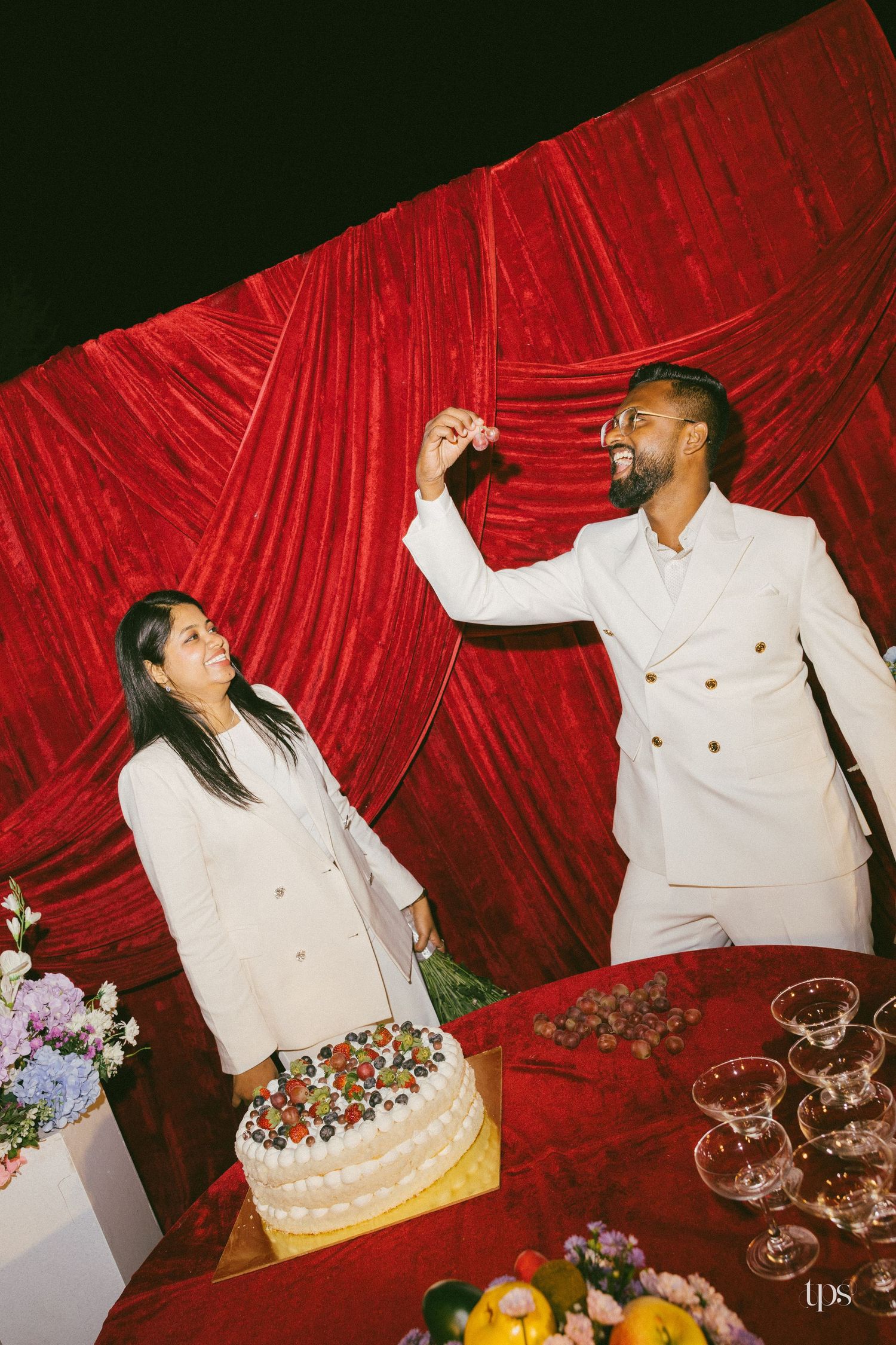 Couple in matching white suits cut and share a decorated berry cake at a wedding reception against a red velvet curtain backdrop.