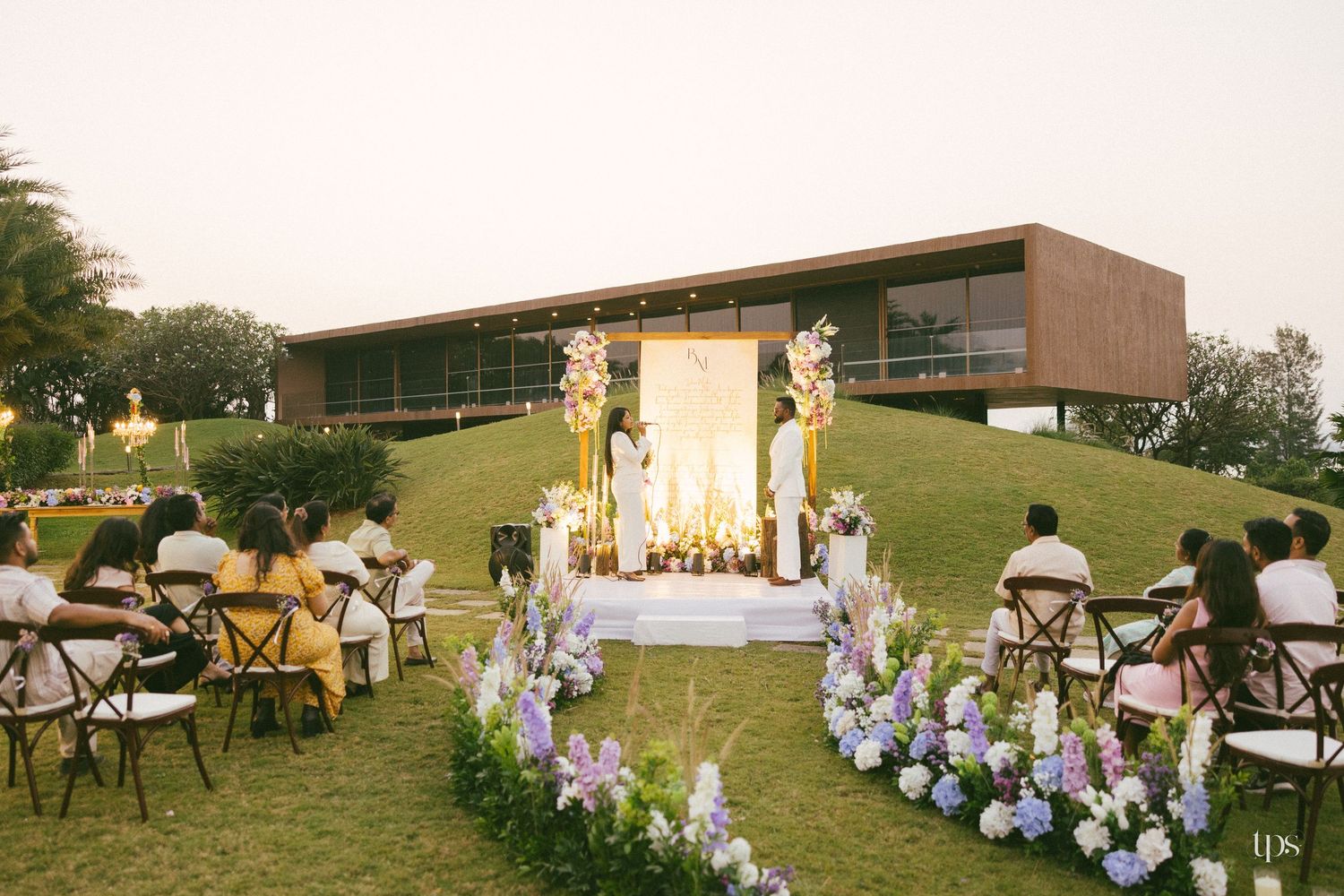 Outdoor garden wedding ceremony on a grassy hill with guests seated among purple and white floral arrangements near a modern building.