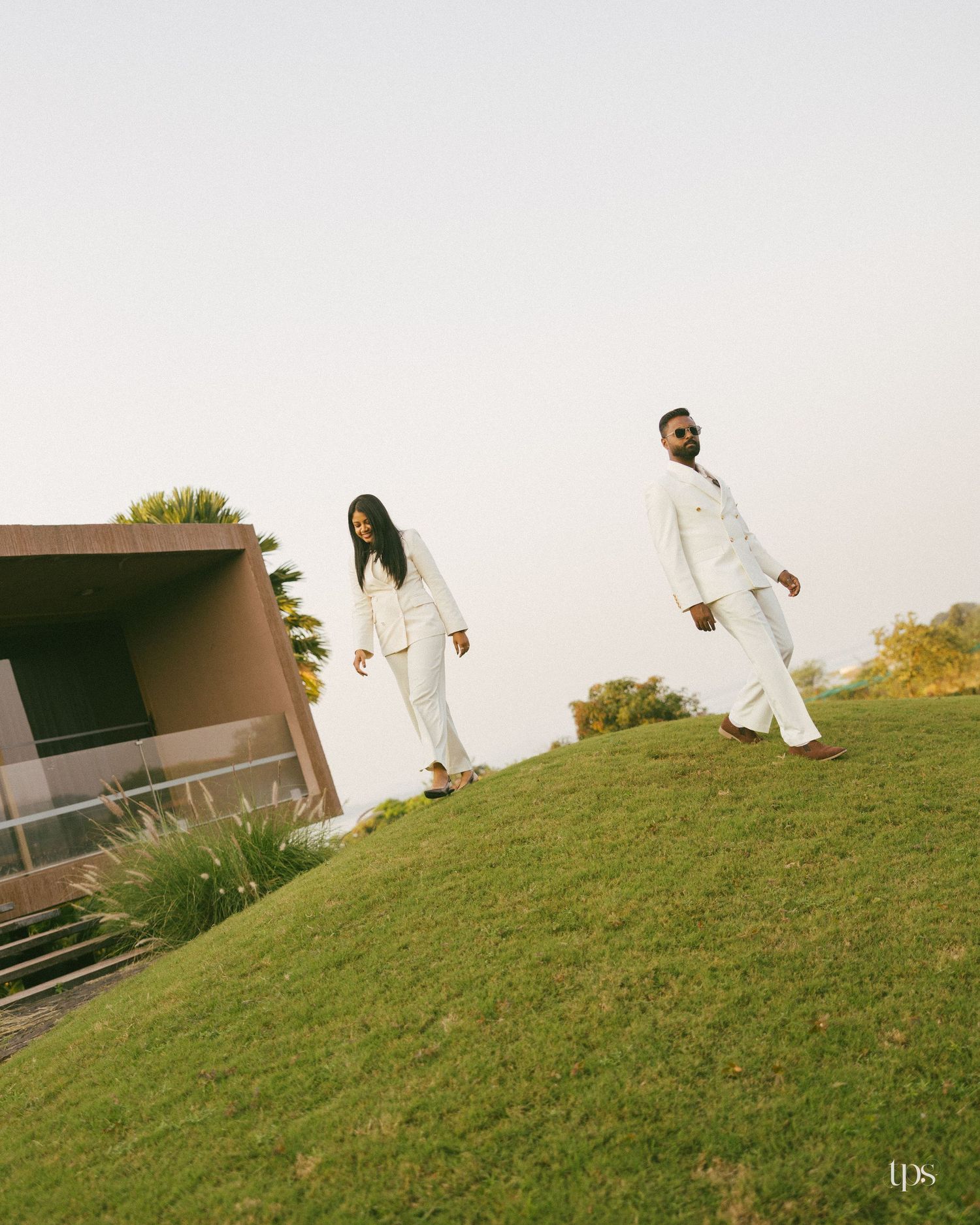 Two people in matching white suits walk down a grassy hillside beside a modern building under a soft, hazy sky.