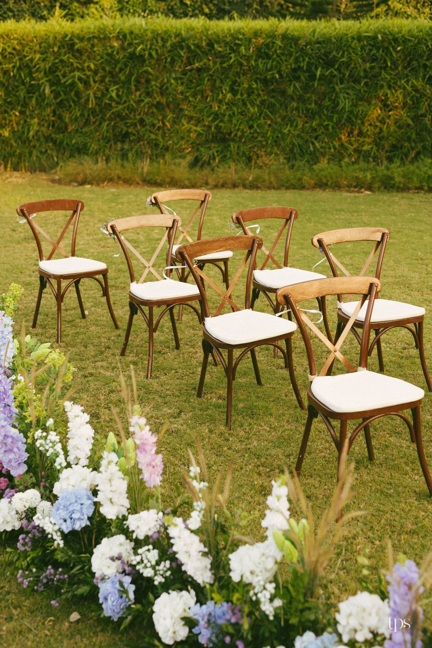 Wooden cross-back chairs with white cushions in staggered rows on a lawn beside lush hydrangea and wildflower arrangements at an outdoor wed