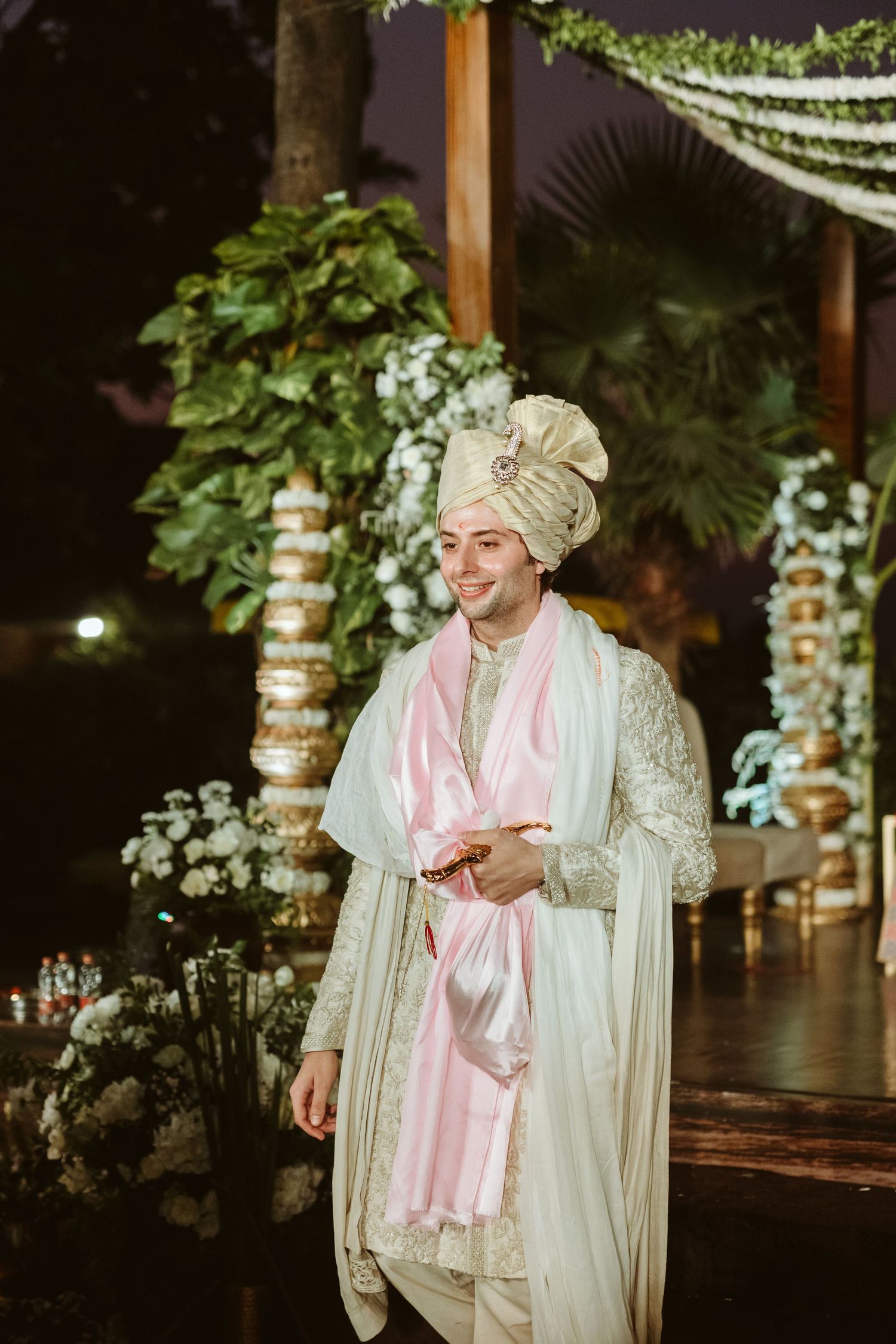 Groom in white sherwani and pink dupatta wearing a cream turban at an elegant outdoor Indian wedding ceremony.