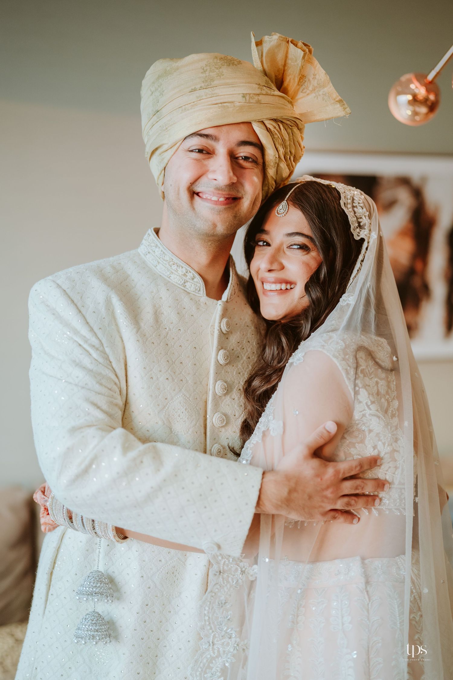 Indian couple in traditional wedding attire, groom in cream sherwani and gold turban embracing bride in white lehenga with veil.