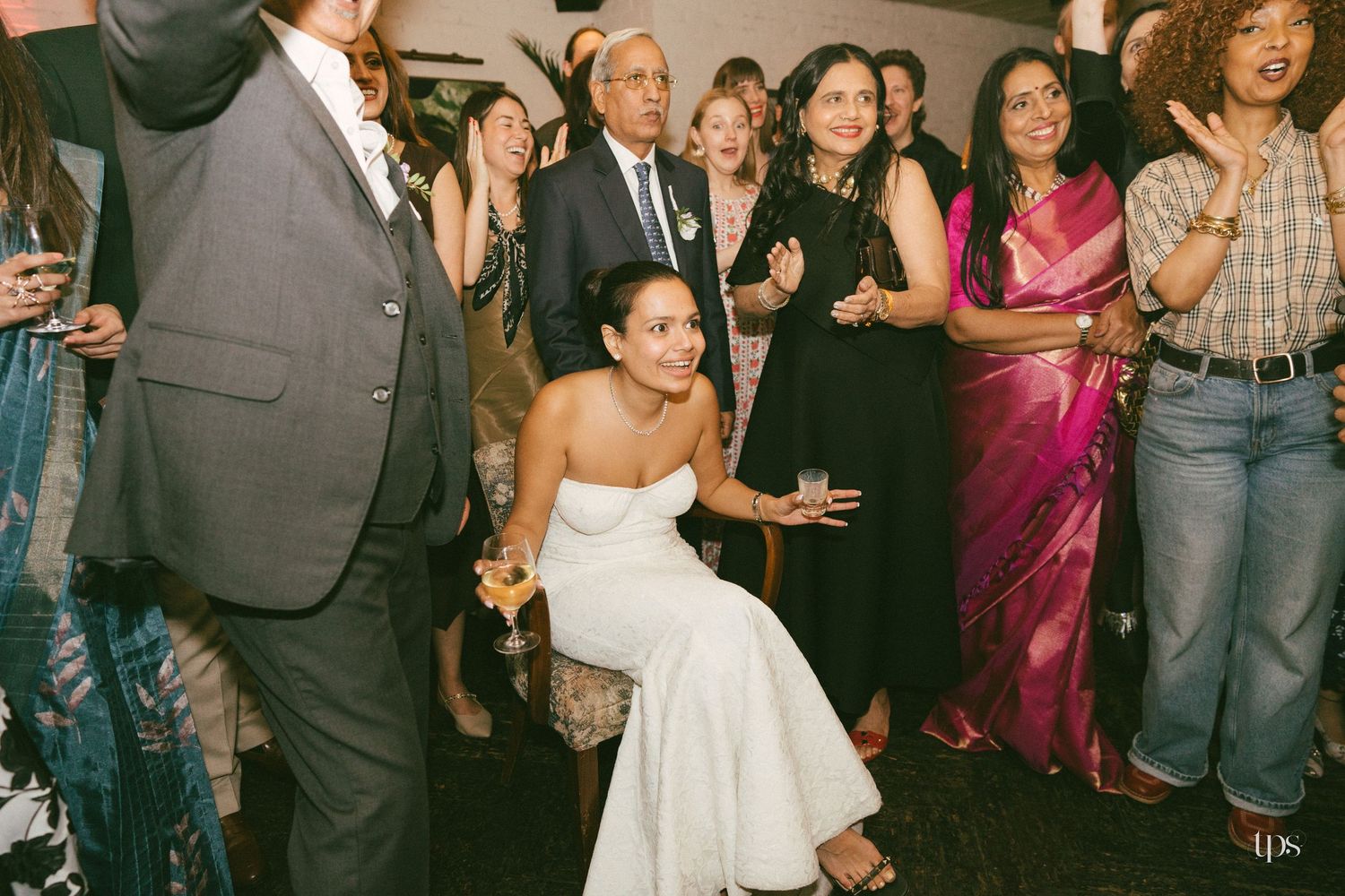Bride in white strapless gown crouches smiling at wedding reception surrounded by guests in formal attire.