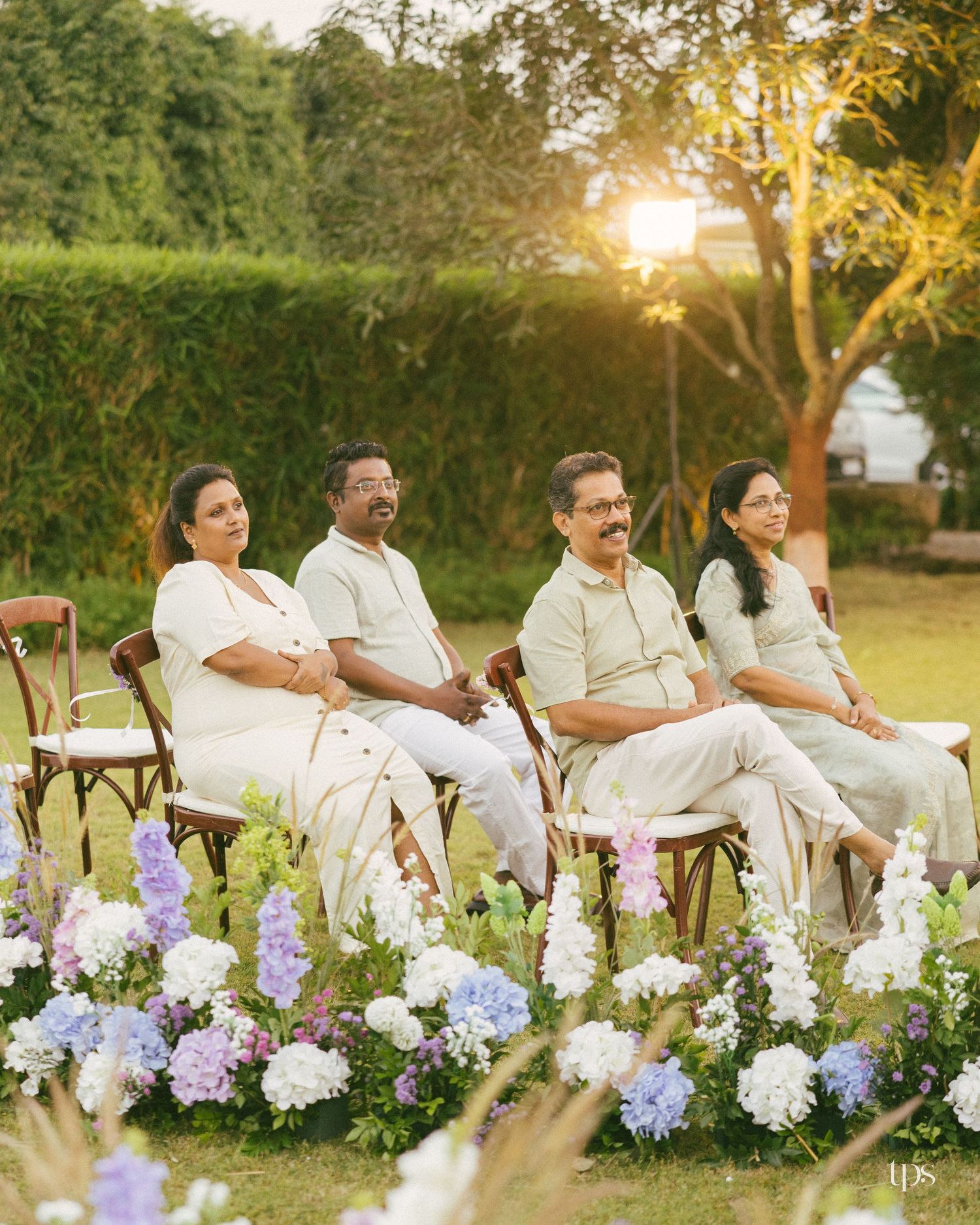 Four people in coordinated cream and sage outfits sit on wooden chairs in a sunlit garden behind arrangements of white and purple flowers.