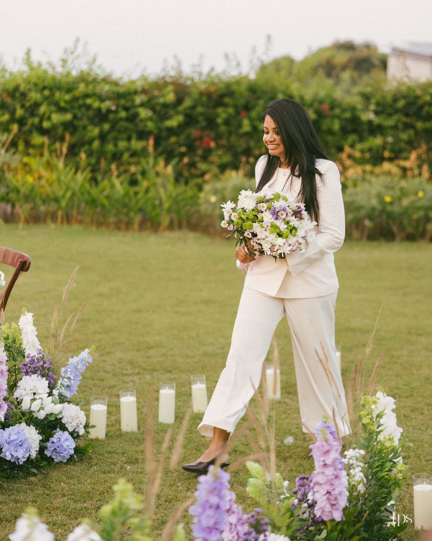 Smiling bride in a white pantsuit carries a lavender and white bouquet down a grass aisle lined with candles and flowers.