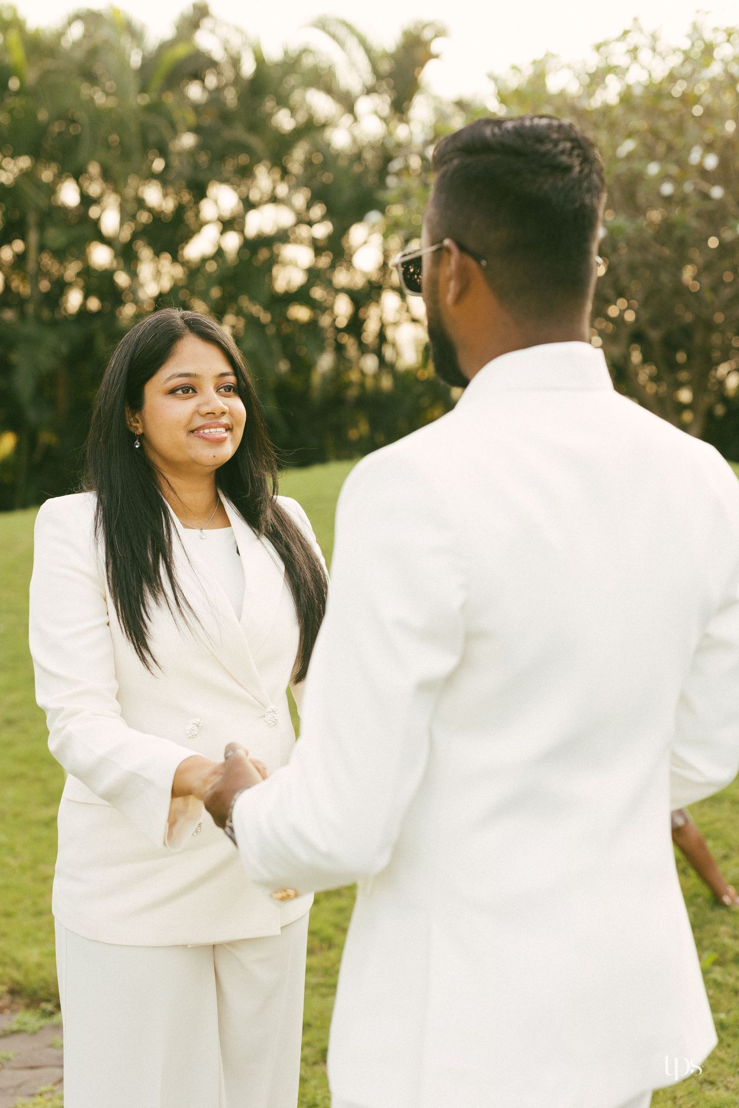 Smiling woman and man in matching white suits holding hands outdoors on a sunlit green lawn with tall trees in the background.