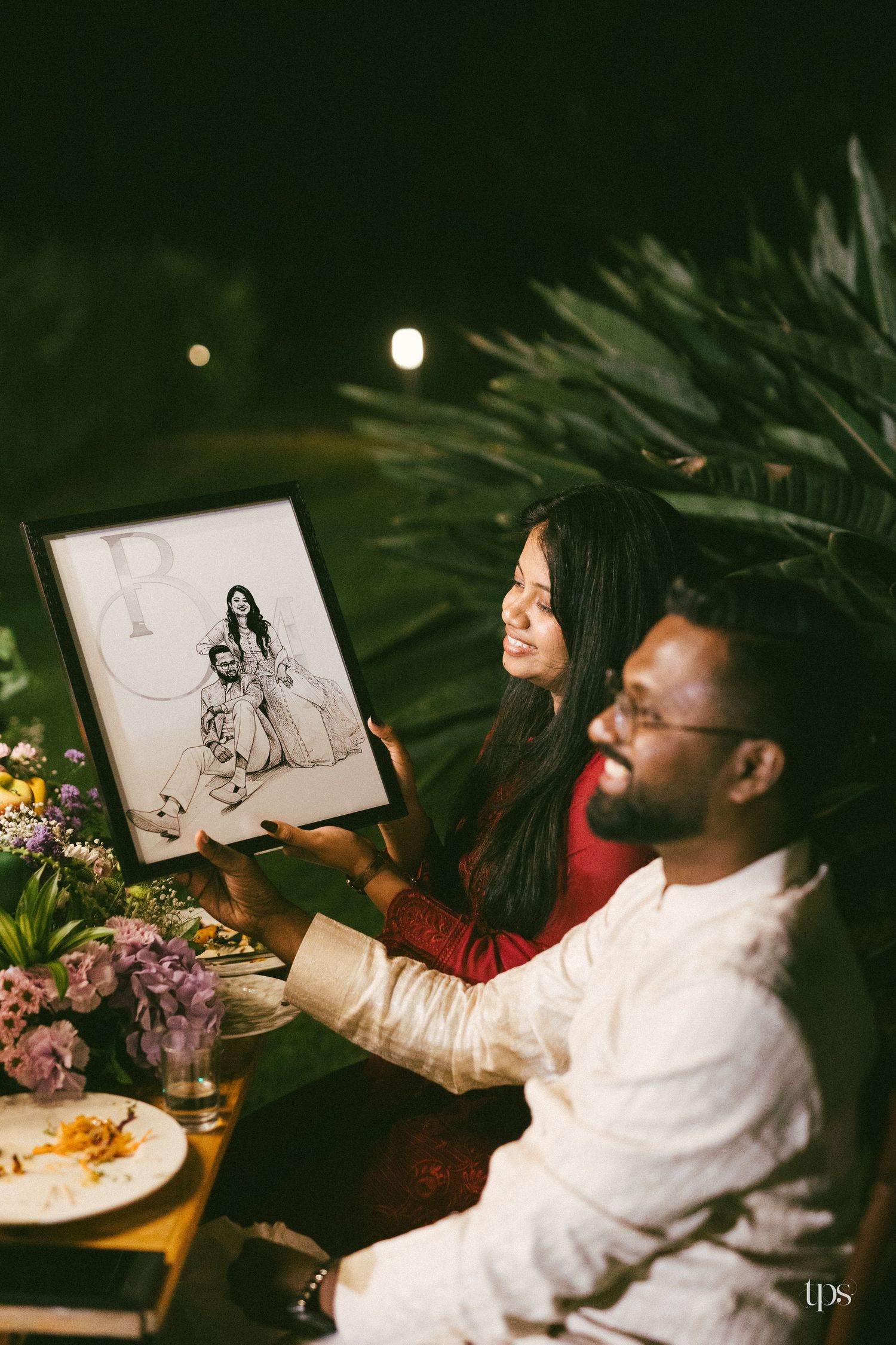 A smiling couple holds a framed black-and-white illustrated portrait at an outdoor evening dinner table with flowers and plates.
