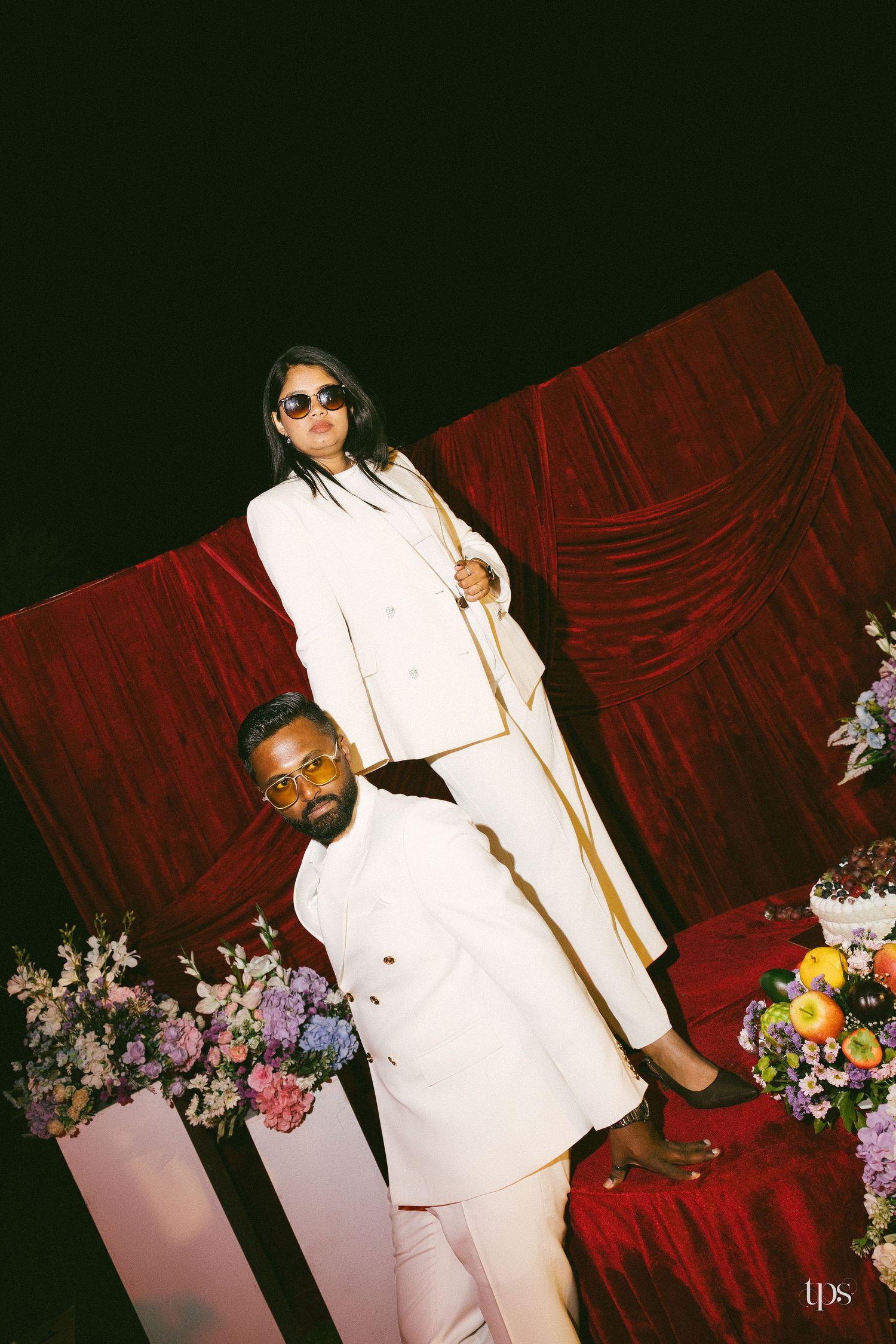 Two people in matching cream suits and sunglasses pose near red velvet drapes, with pastel flower arrangements and fresh fruit nearby.