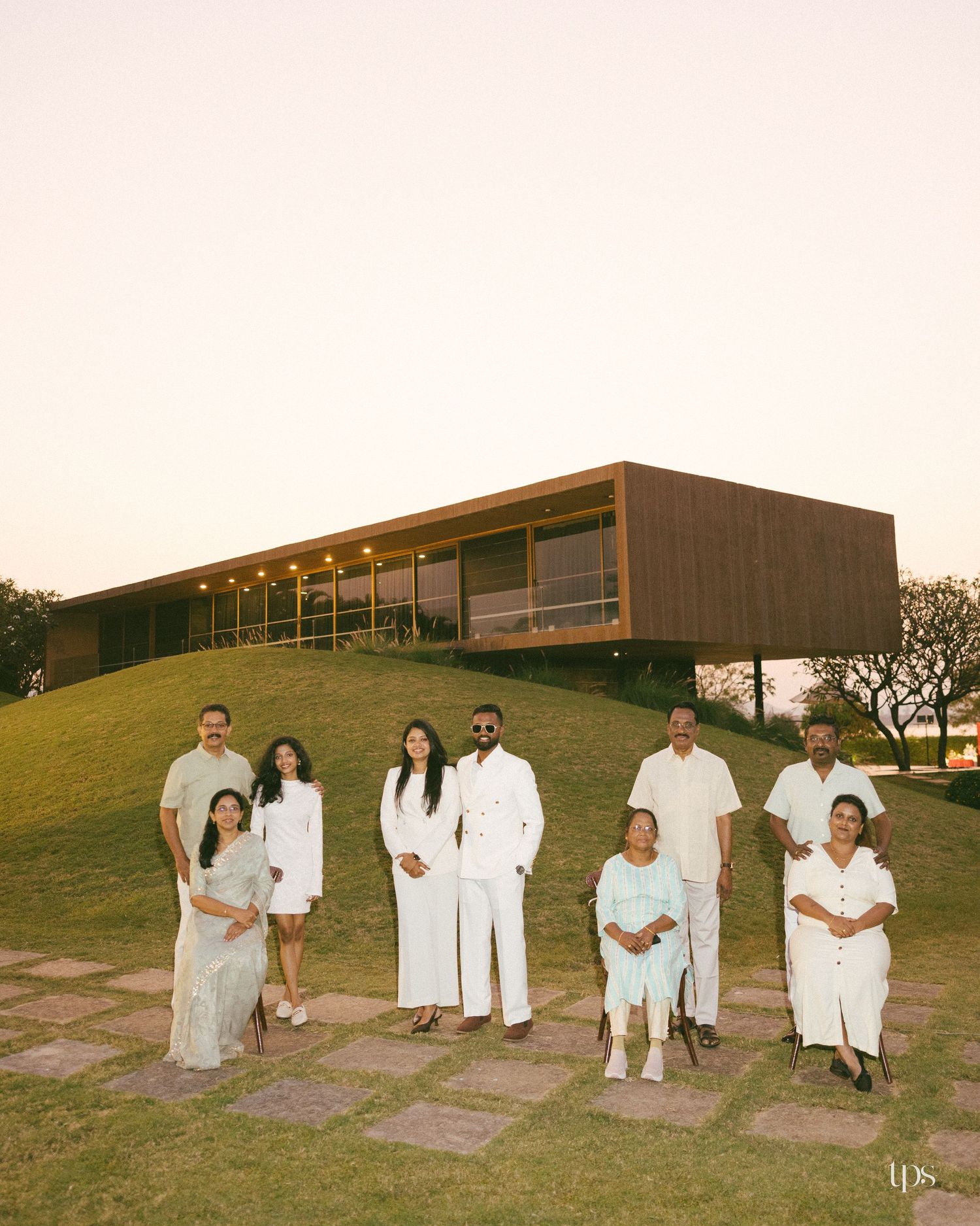 Family dressed in white and pastel outfits poses on a grassy hillside before a modern wood-and-glass house at sunset.