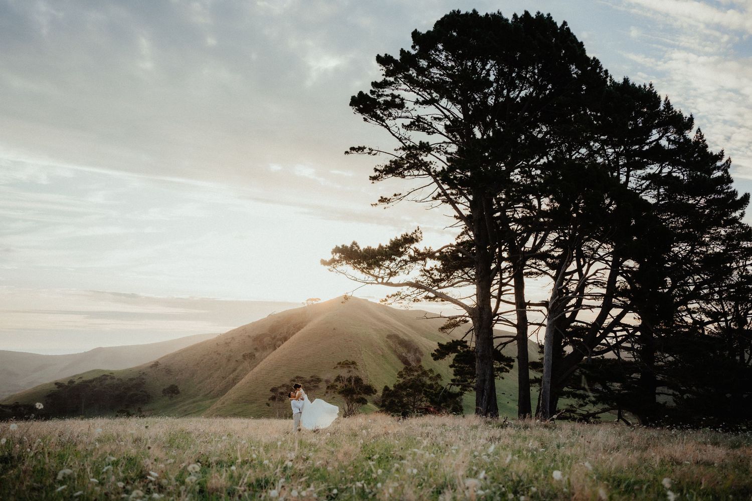 Bride in a white dress stands in a wildflower meadow beneath tall trees with rolling hills at sunset.