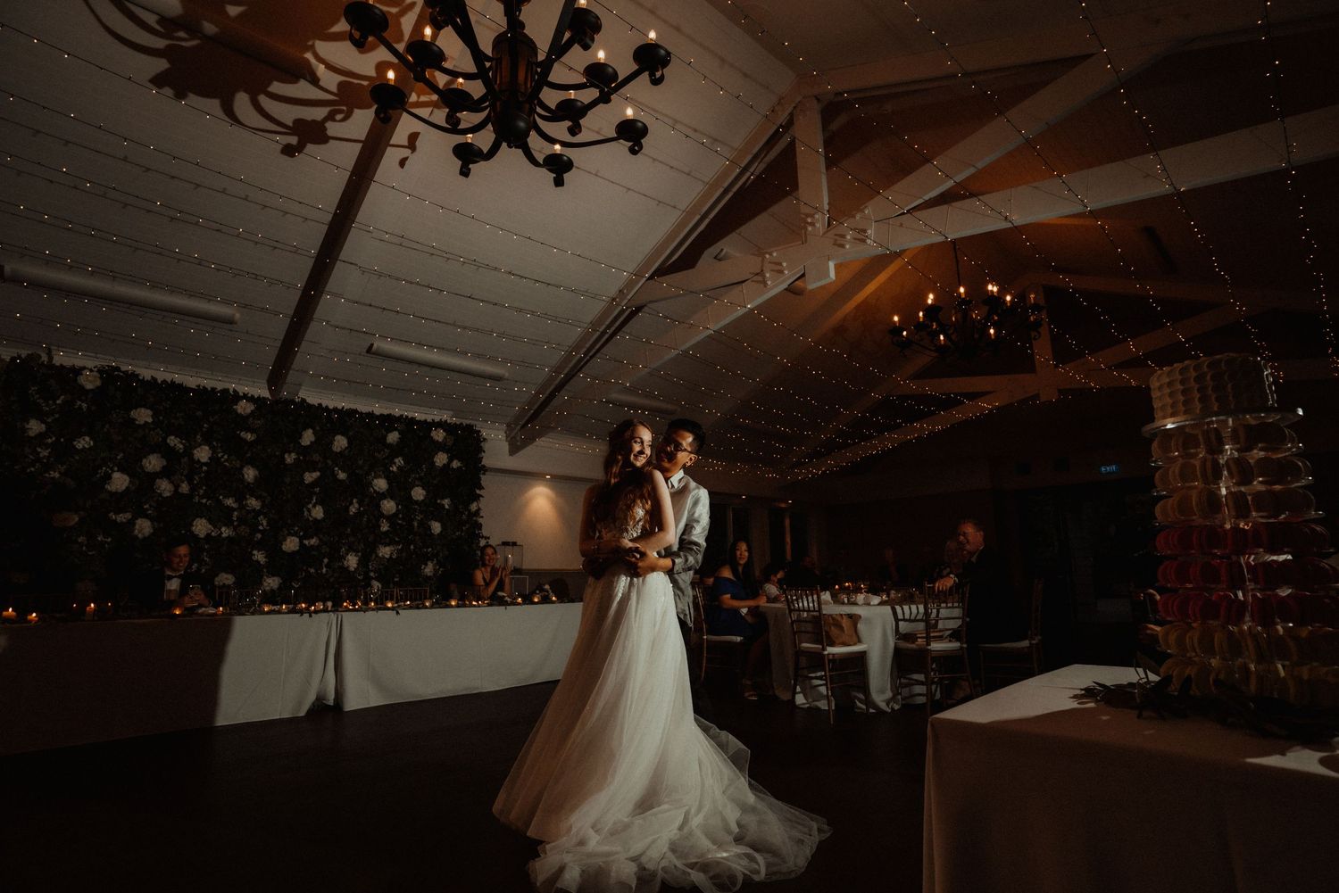 Bride and groom share a romantic first dance in a dimly lit reception hall with a chandelier and champagne tower.