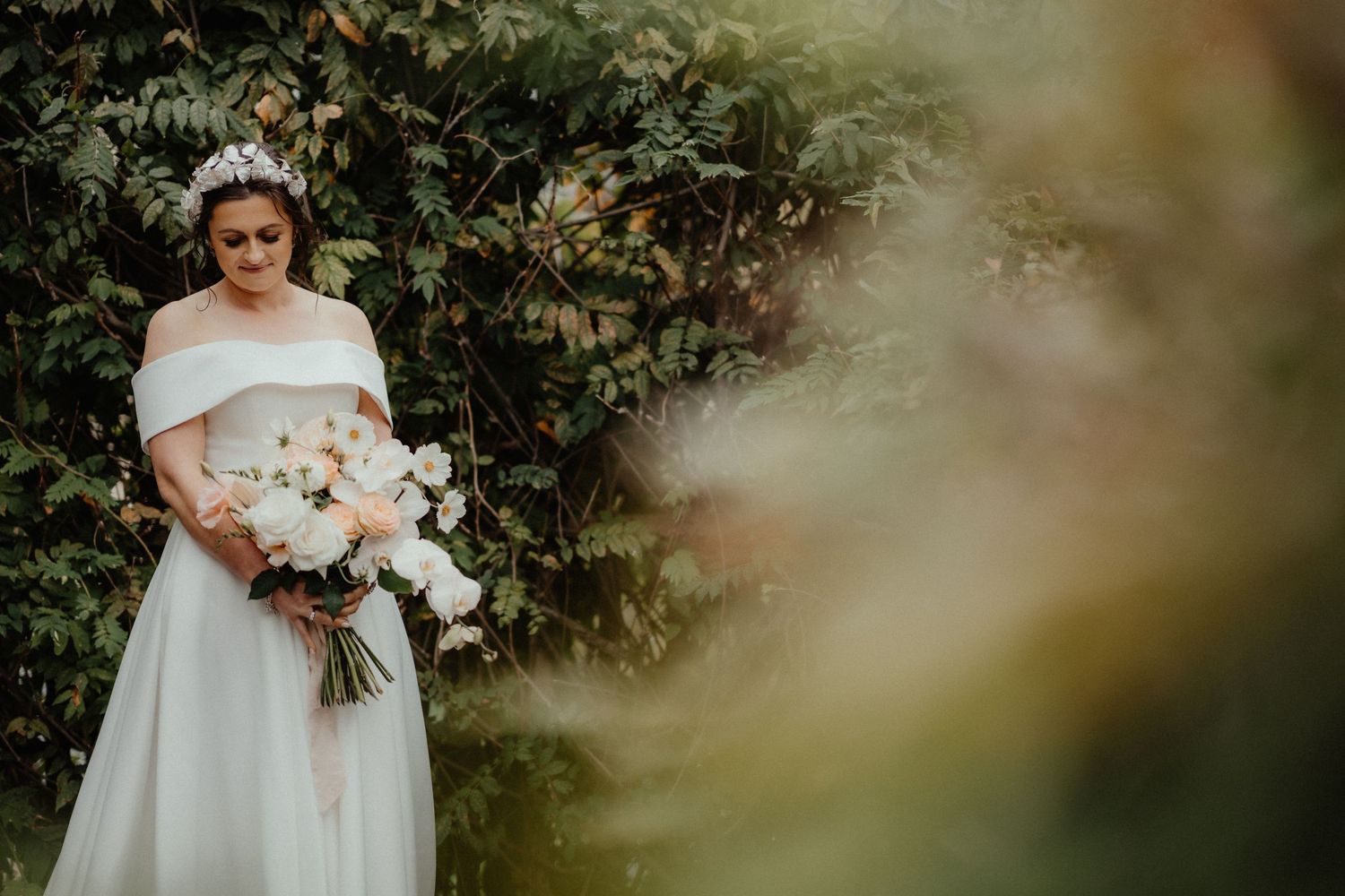 Bride in an off-shoulder white gown and tiara holds a white and blush bouquet in front of lush green foliage.