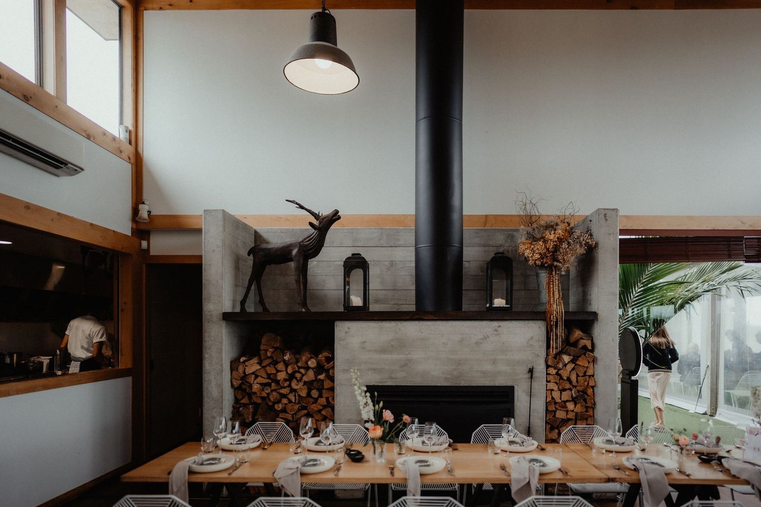 Rustic modern wedding reception room with a long wooden table, clear chairs, a concrete fireplace, stacked firewood and a deer sculpture.