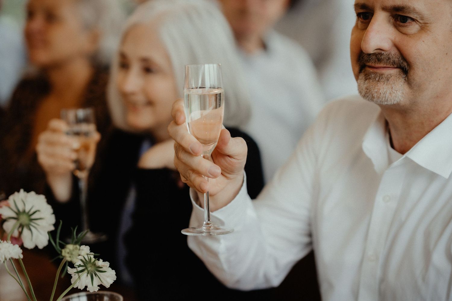 A smiling older man raises a glass of champagne at a wedding reception while guests sit behind him and white flowers rest in the foreground.