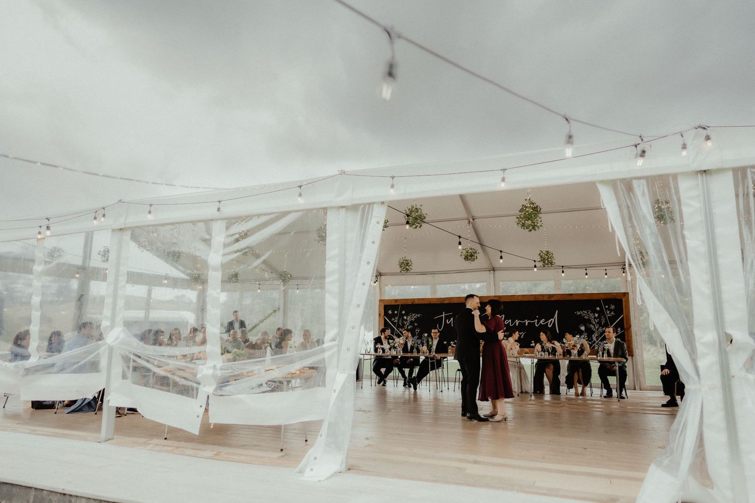 Newlyweds share a first dance on a wooden floor inside a clear marquee tent as wedding guests look on.
