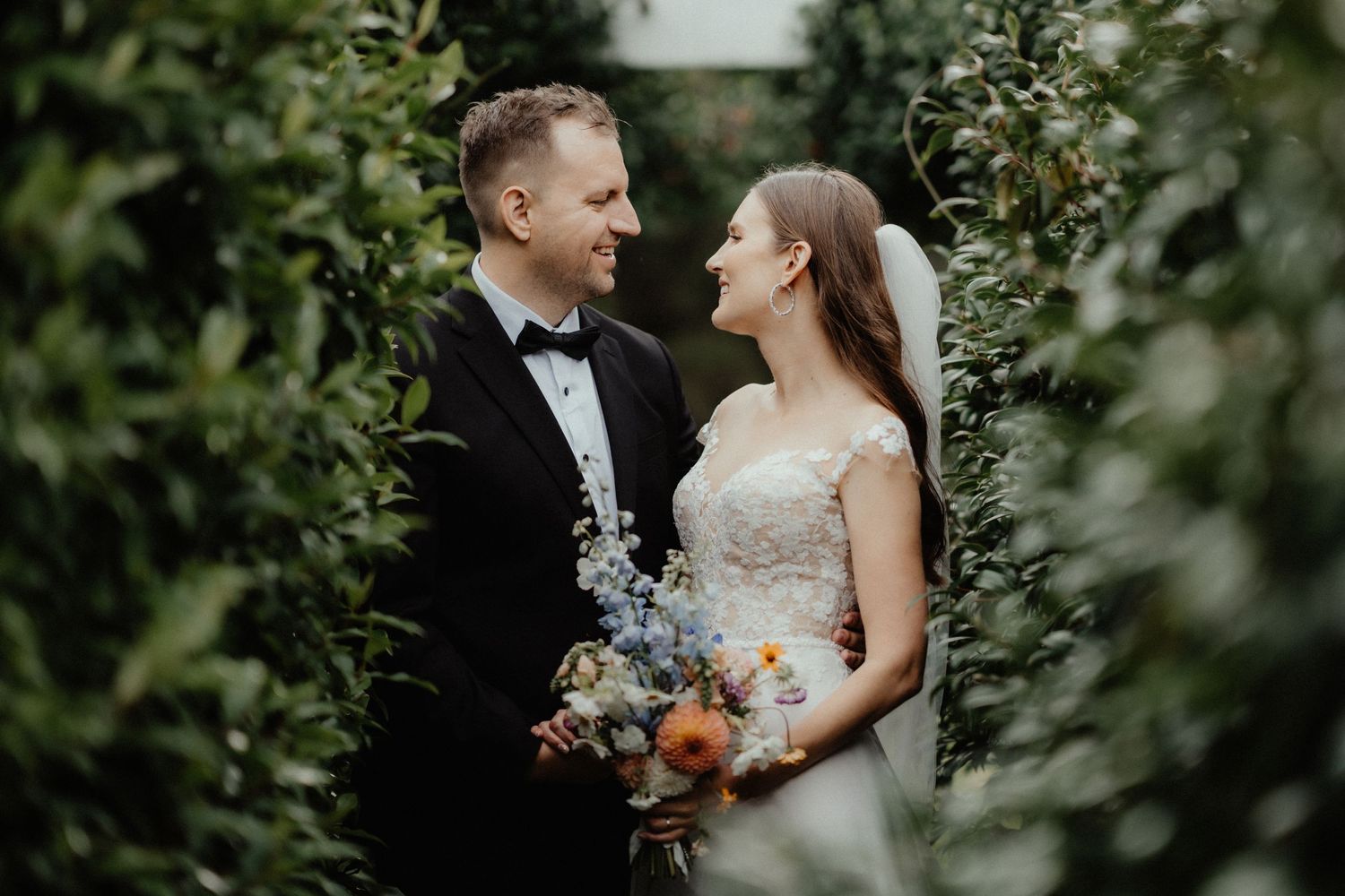 Bride and groom smile at each other between lush green hedges, holding a colorful bouquet of wildflowers.