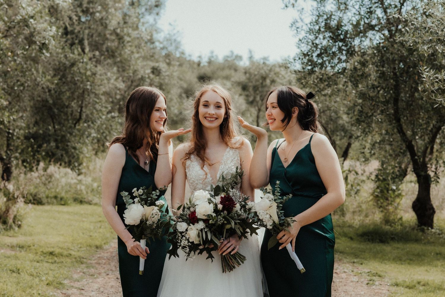 Bride in a white gown laughs with two bridesmaids in emerald green dresses holding bouquets in an olive grove.