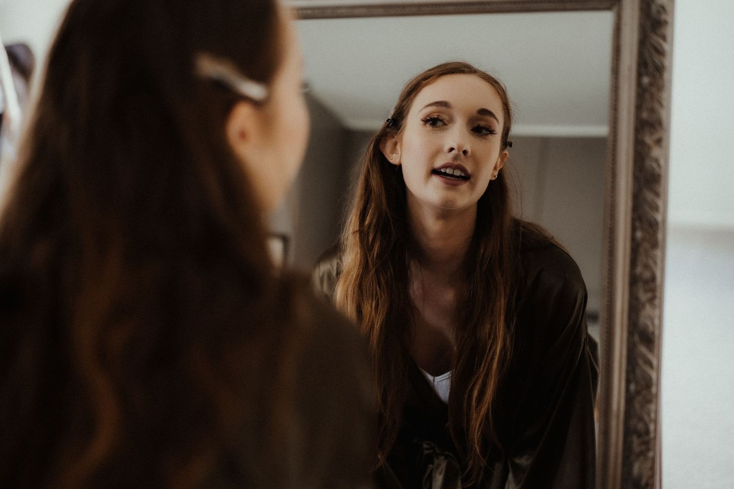 A young woman with long auburn hair speaks while looking at her reflection in a large ornate mirror.