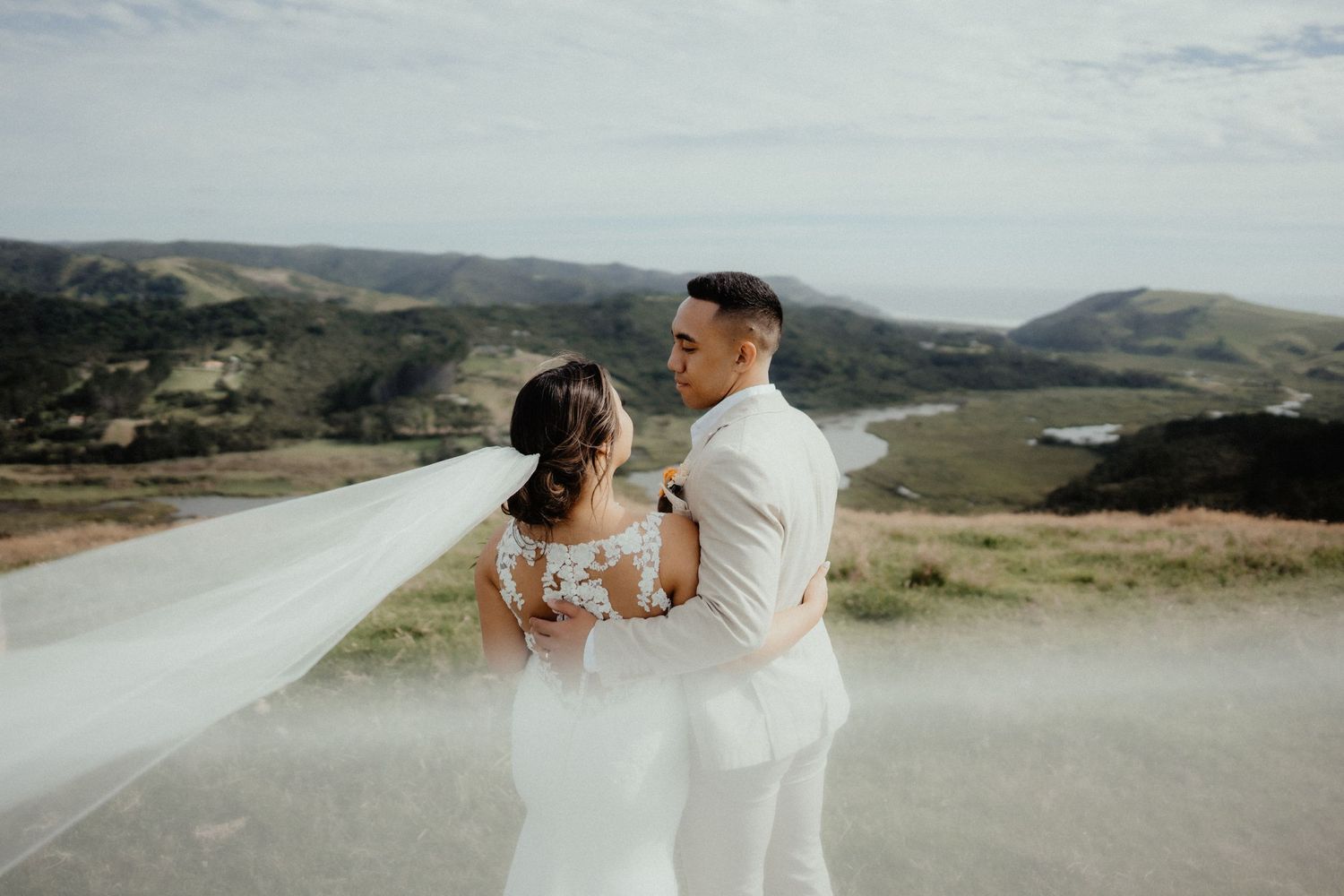 Couple embraces on a hilltop overlooking rolling green hills as the bride's long veil blows in the wind.