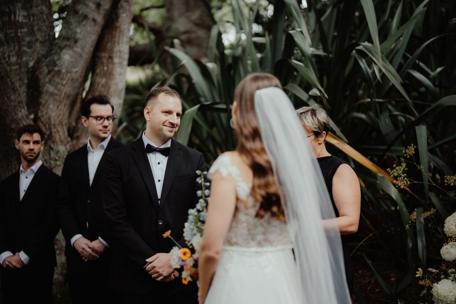 Groom in a black tuxedo and bow tie smiles as groomsmen look on during an outdoor wedding ceremony with the bride in a veil.