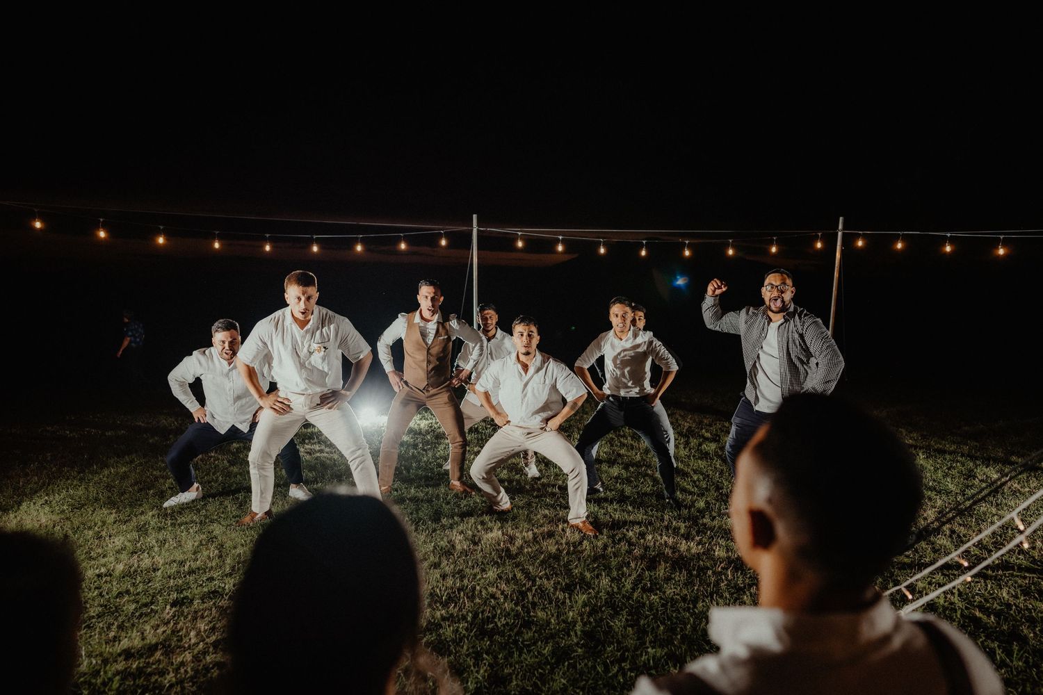 Groomsmen perform an energetic haka dance on the lawn at night beneath glowing festoon string lights.
