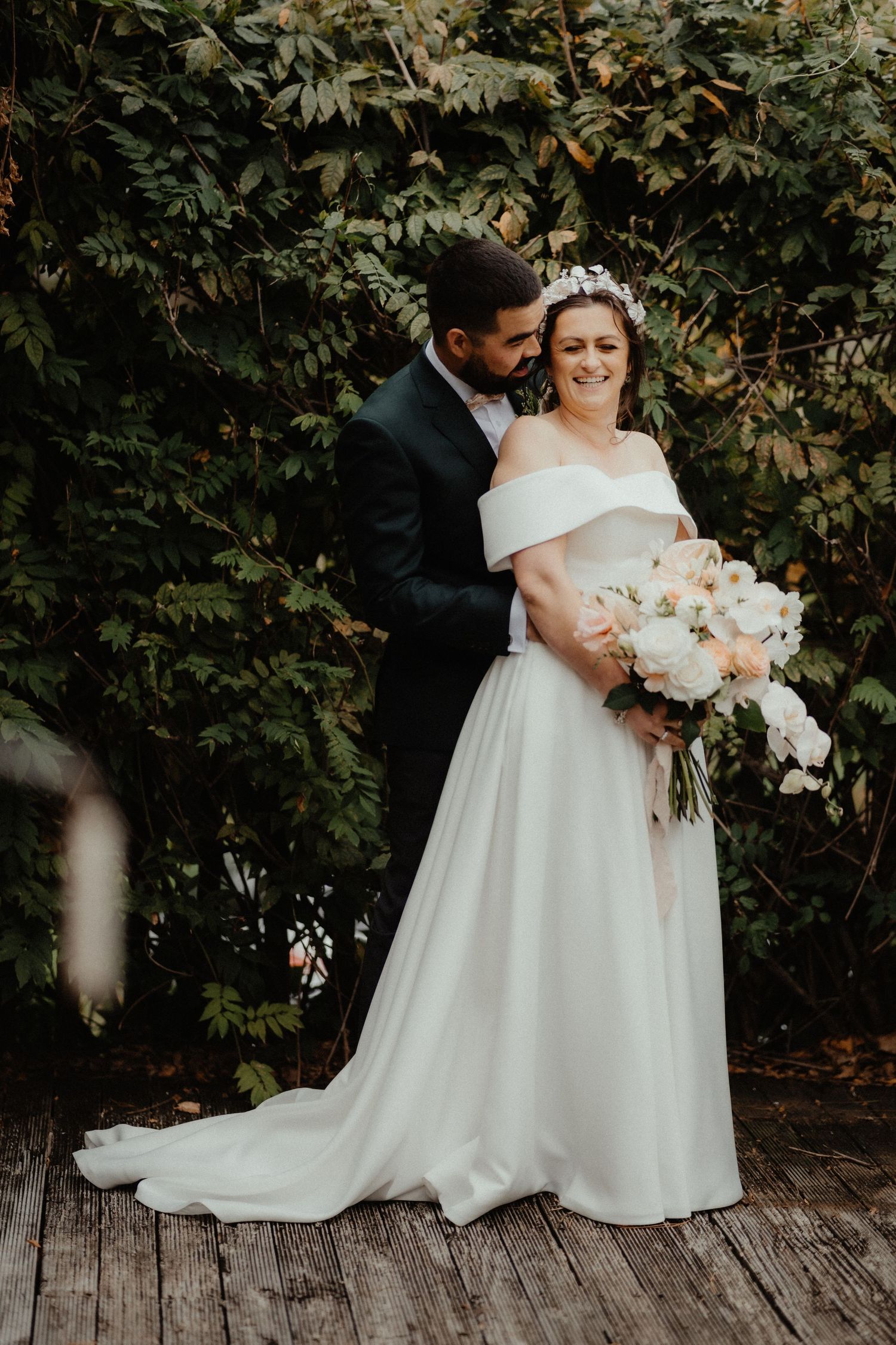 A smiling bride in an off-shoulder white gown holds a bouquet while her groom hugs her from behind in front of a leafy hedge.