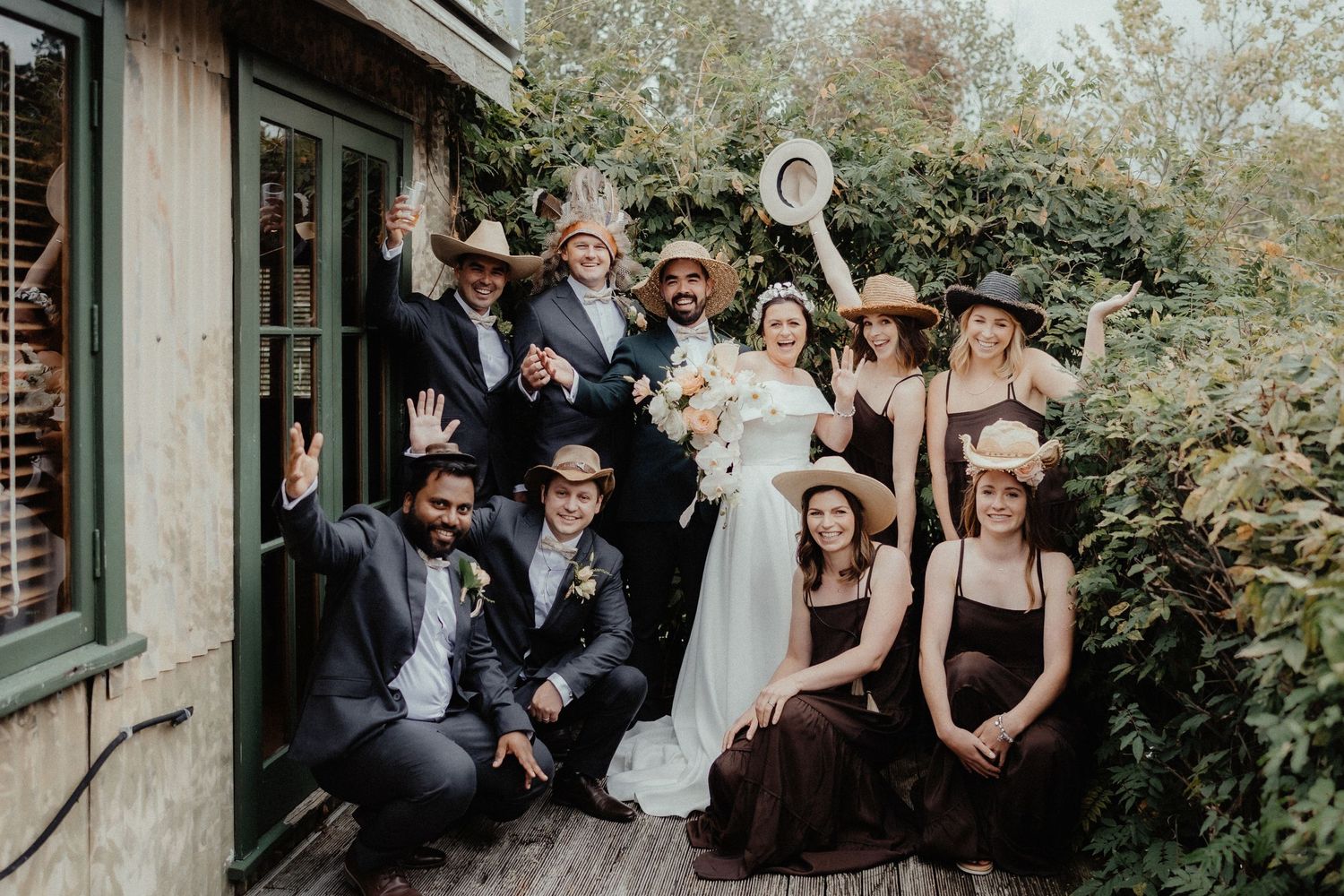 Bride and groom pose with joyful bridesmaids and groomsmen in rustic hats, cheering outside a vine-covered building.
